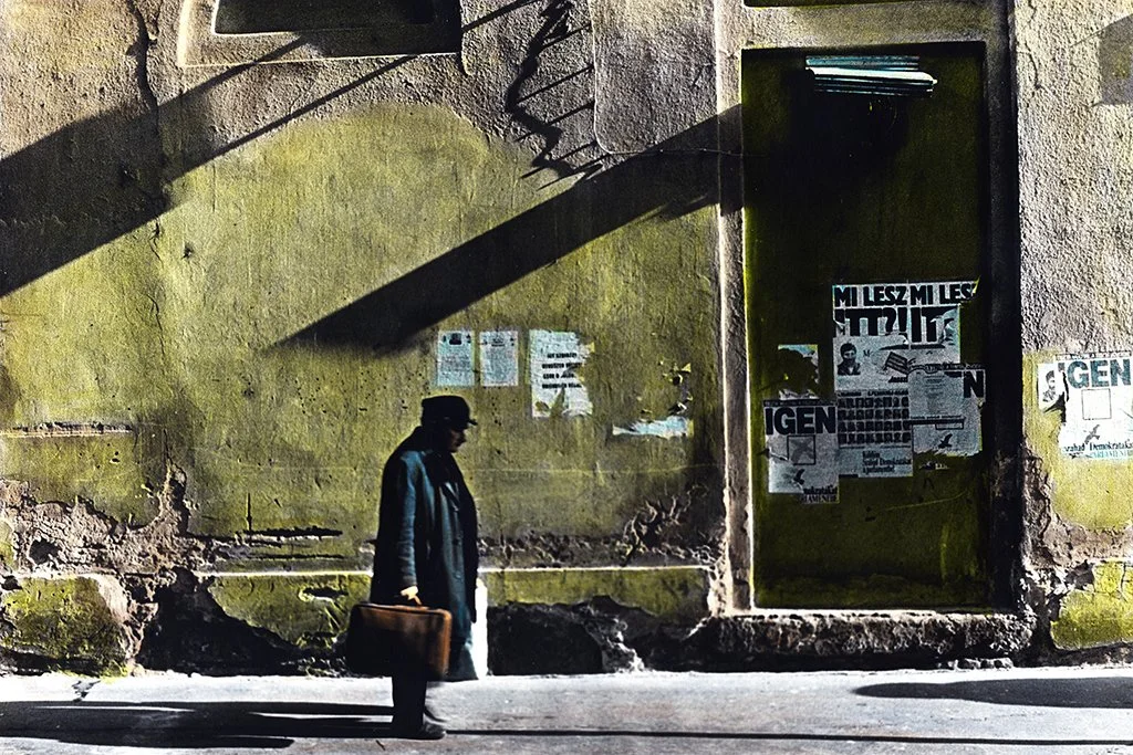 A man wearing a hat and coat, carrying a brown briefcase, walking past an old, weathered yellow wall with posters and a square window, casting shadows on the wall.
