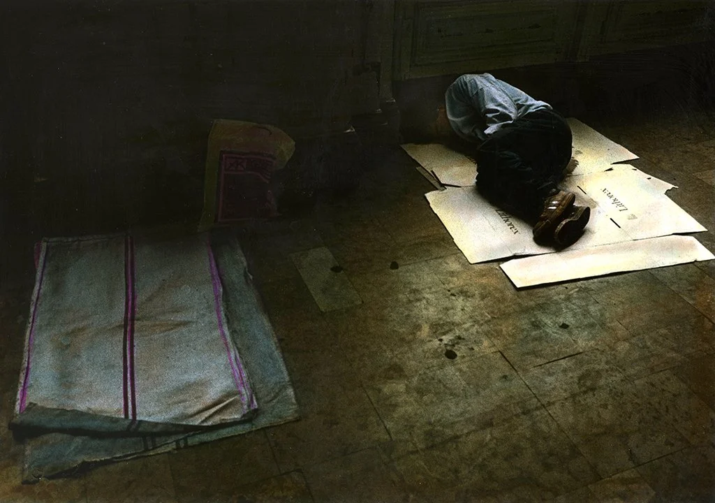Child kneeling on the floor, reading a piece of paper in a dimly lit room, with a bag and a box nearby.