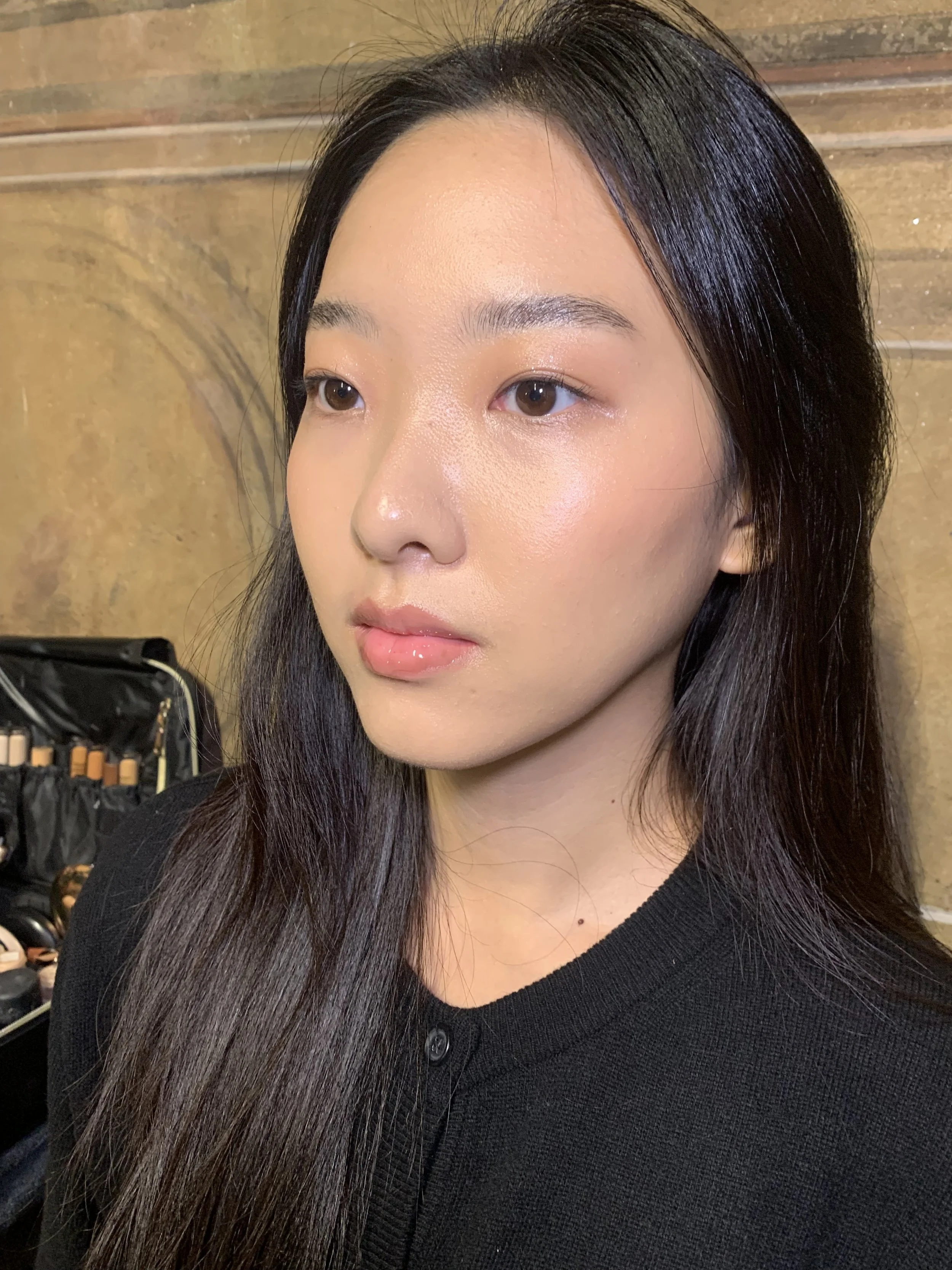 Close-up of a young woman with long black hair, wearing a black top, standing in front of a wooden background.