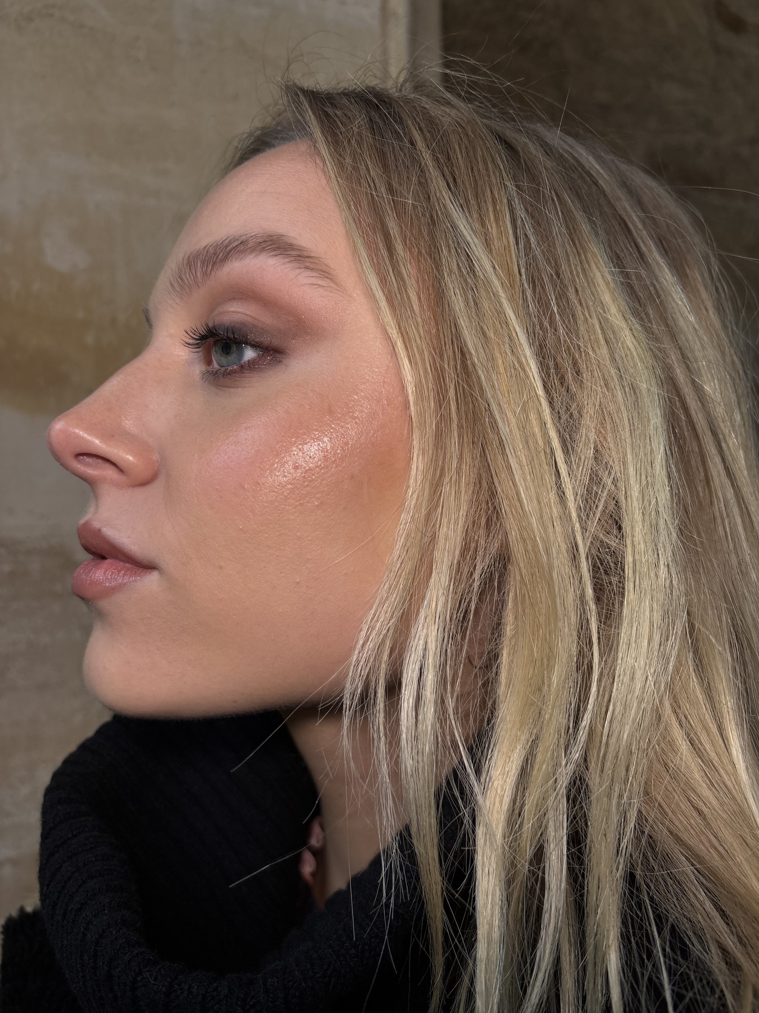 Close-up side profile of a woman with blonde hair, light makeup, and a black turtleneck sweater against a beige background.