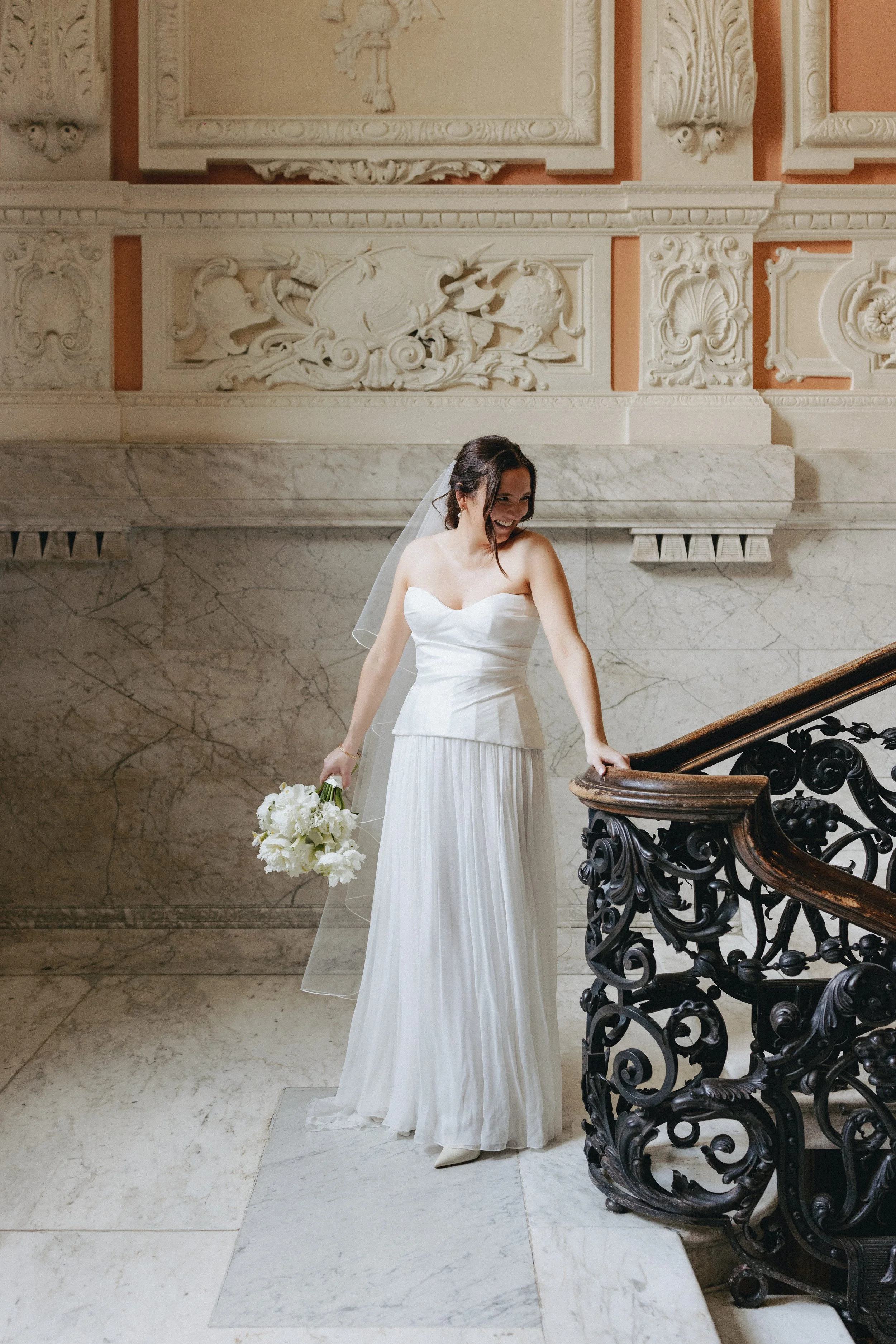 A bride in a strapless white wedding dress holding a bouquet of white flowers, standing on a marble staircase with ornate sculpted wall decor in the background, smiling and looking down.