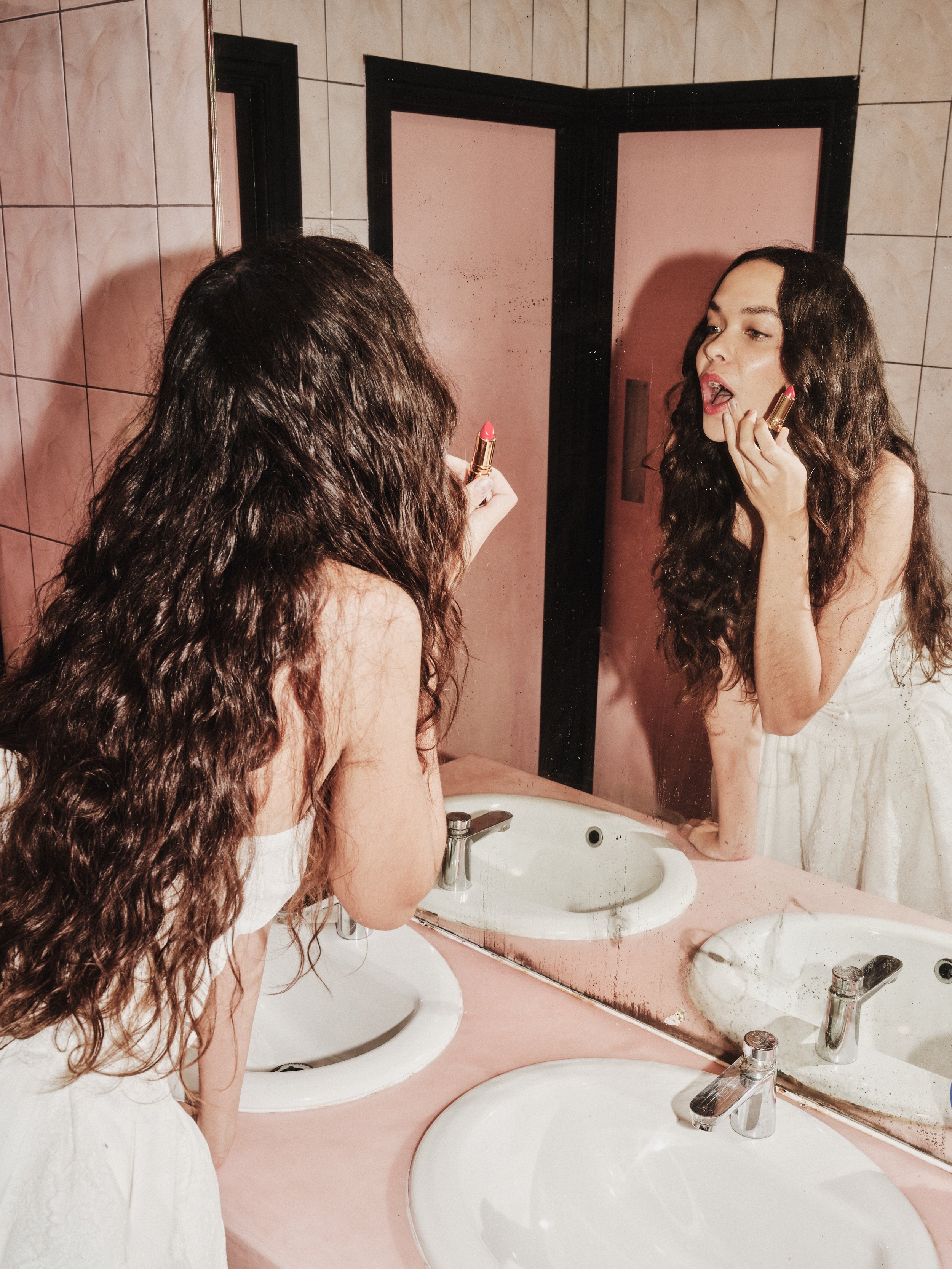 A woman with long curly hair applying pink lipstick in front of a bathroom mirror.