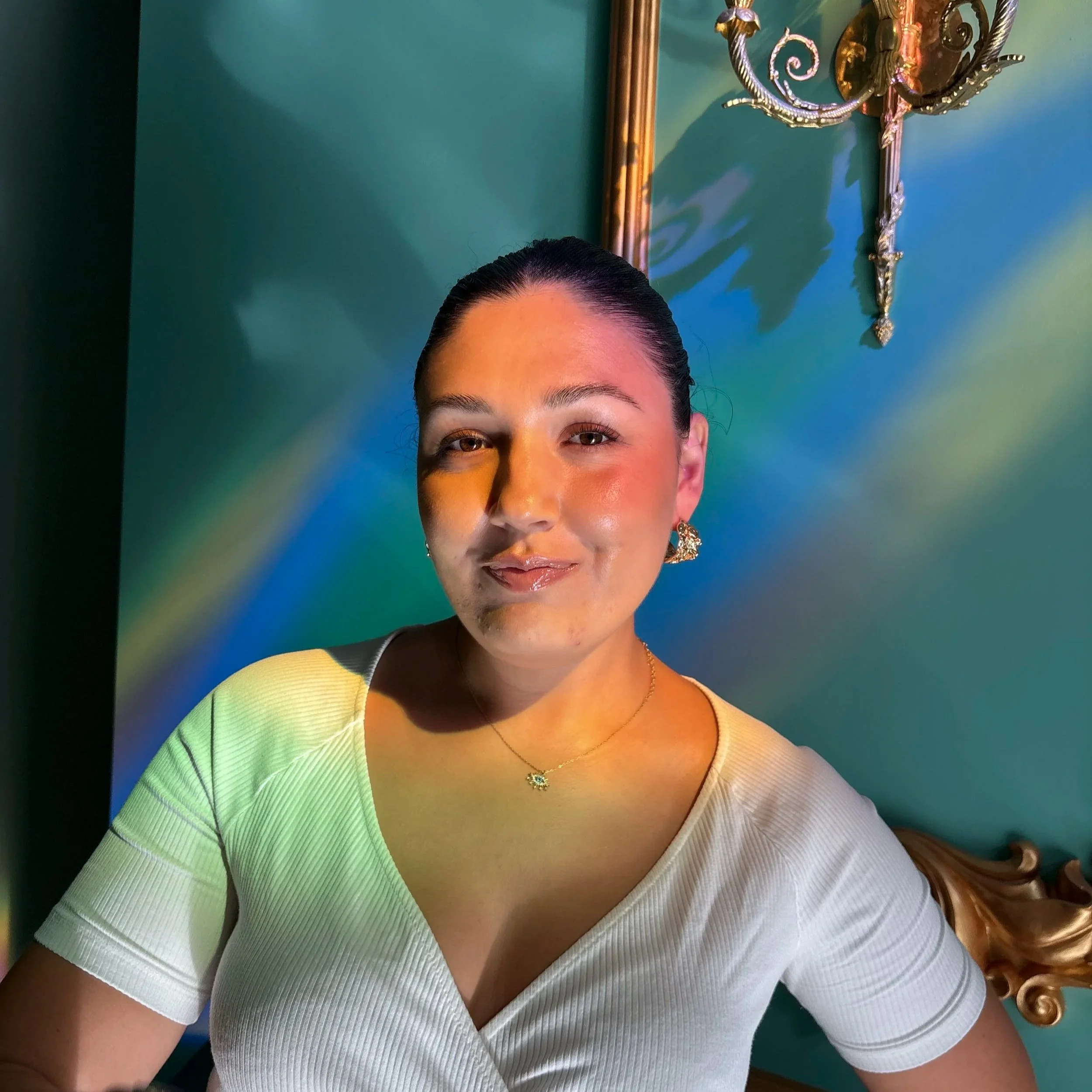 A woman with dark hair pulled back, wearing a white top, gold jewelry, and sitting against a colorful background with a golden framed mirror and ornate wall sconce.