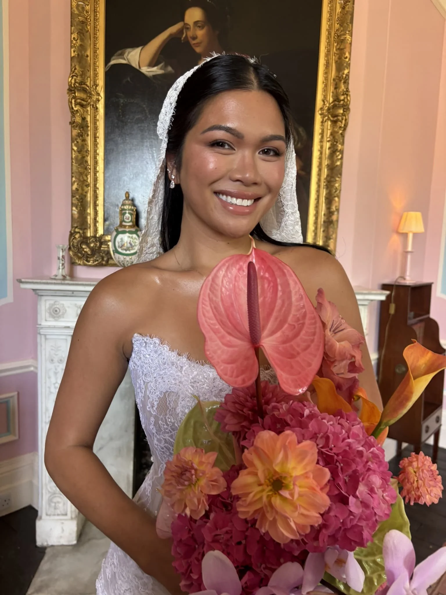 A woman in a white dress with lace details smiles while holding a bouquet of pink, orange, and purple flowers. She has dark hair and is wearing pearl earrings. Behind her is a large framed portrait of a woman resting her head on her hand, with a gold
