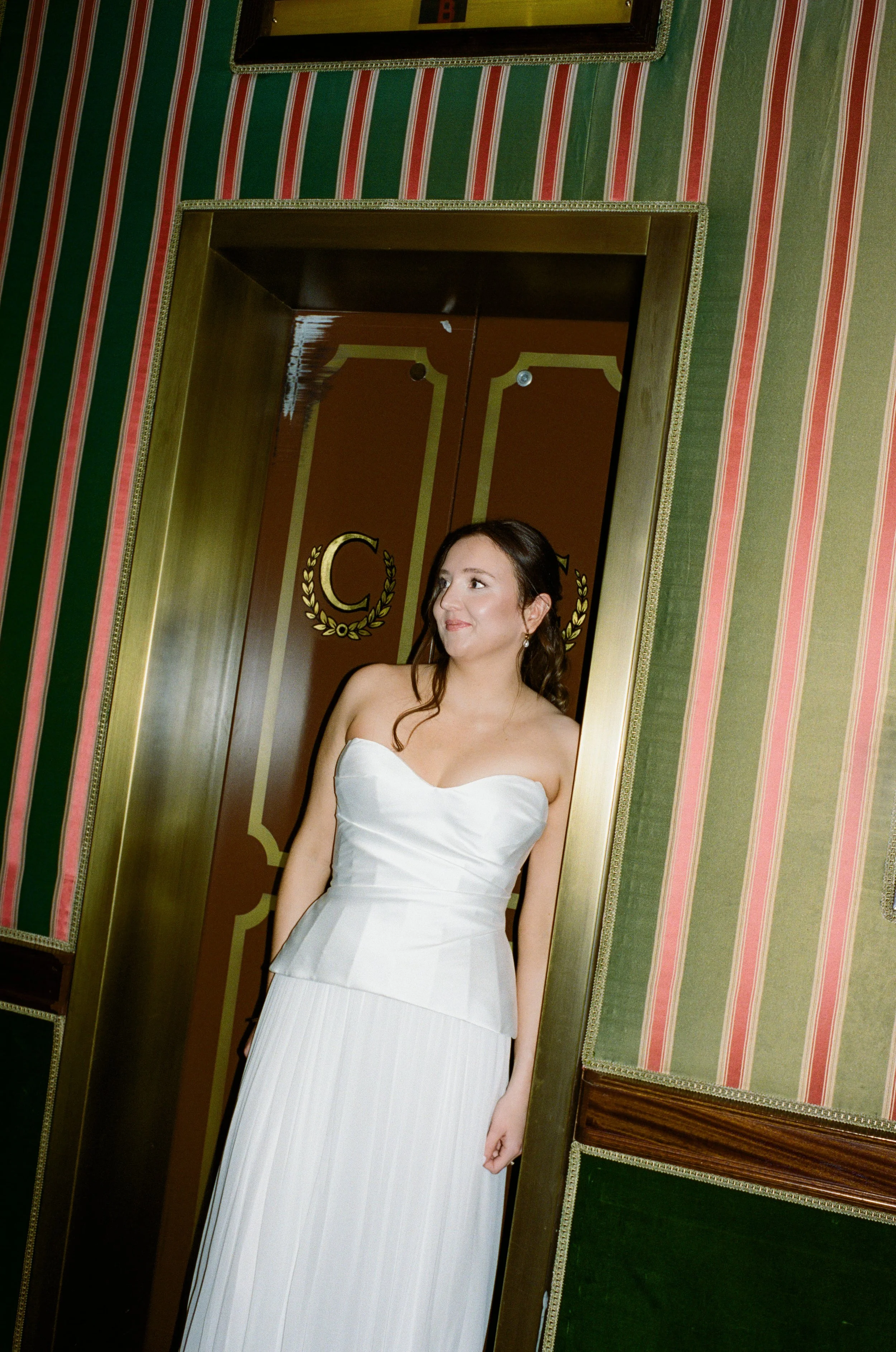 A woman in a strapless white dress standing in front of a large mirror with a gold frame, inside a room with green and red striped wallpaper and ornate gold accents.