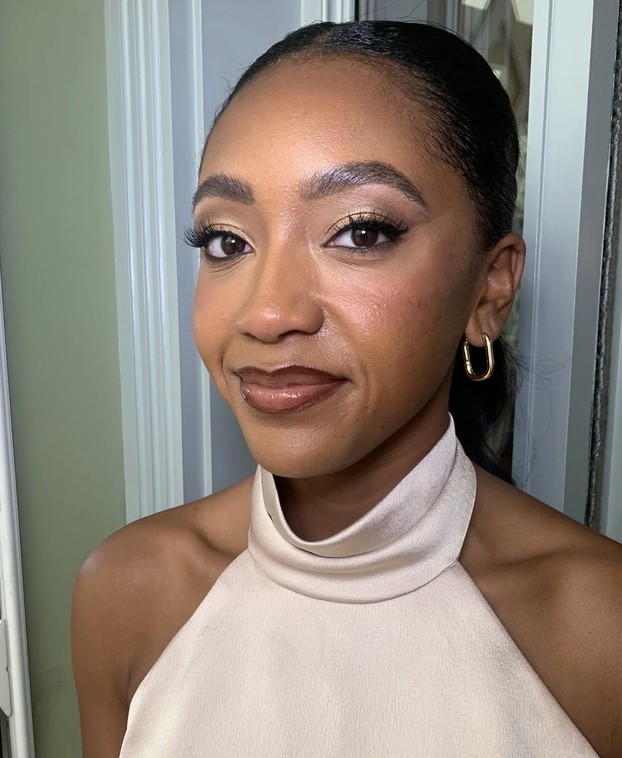 A woman with dark hair tied back, wearing gold hoop earrings and a beige sleeveless turtleneck top, smiling slightly at the camera in front of a white door and green wall.