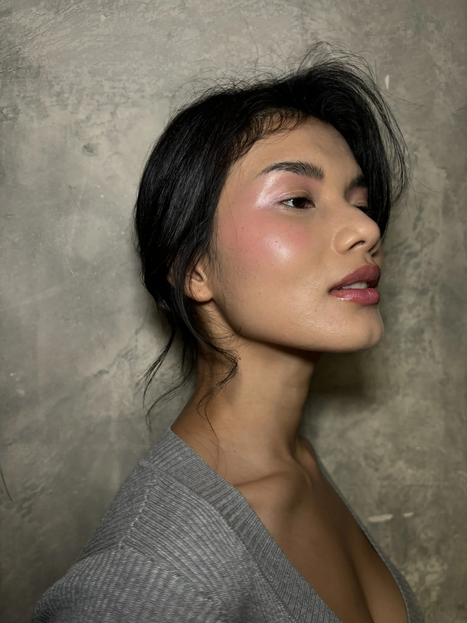 A side profile of a woman with short black hair, wearing a gray shirt, standing against a textured gray wall.