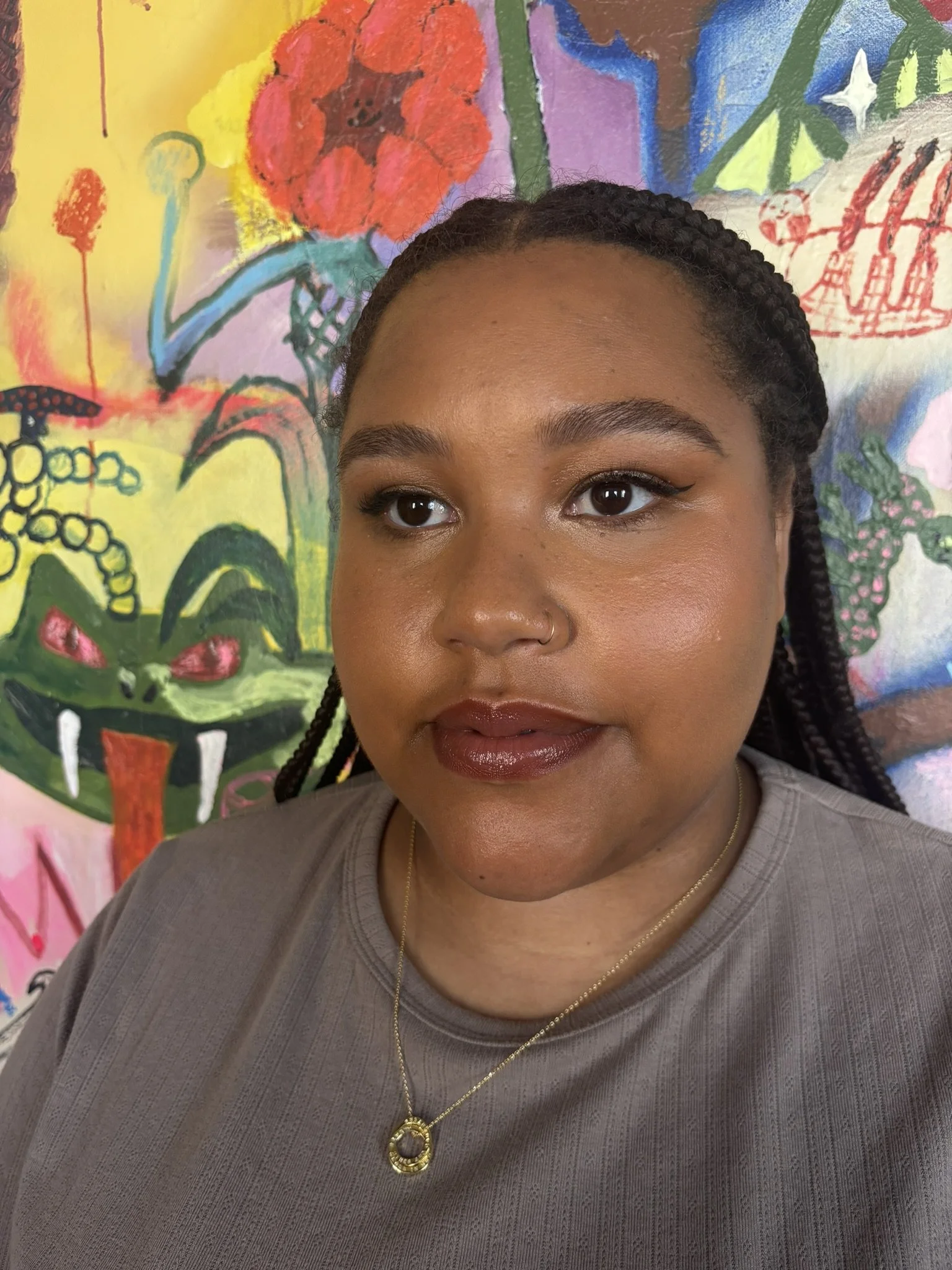 A woman with braided hair and a nose ring, wearing a gray shirt and gold necklace, stands in front of a colorful, abstract mural featuring flowers and a face with red eyes.