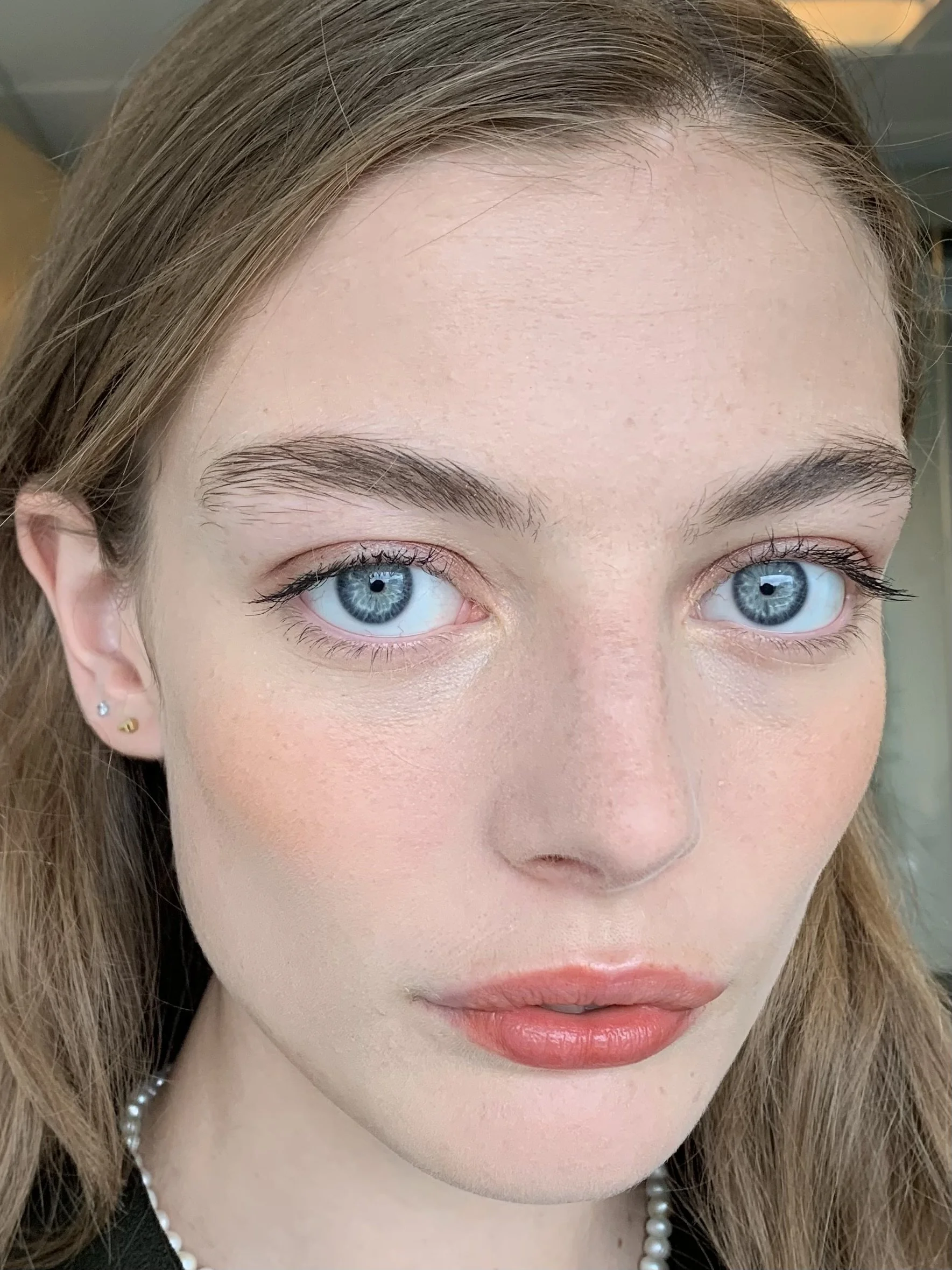 Close-up photo of a woman with light skin, blue eyes, light brown hair, wearing pearl earrings and a pearl necklace.