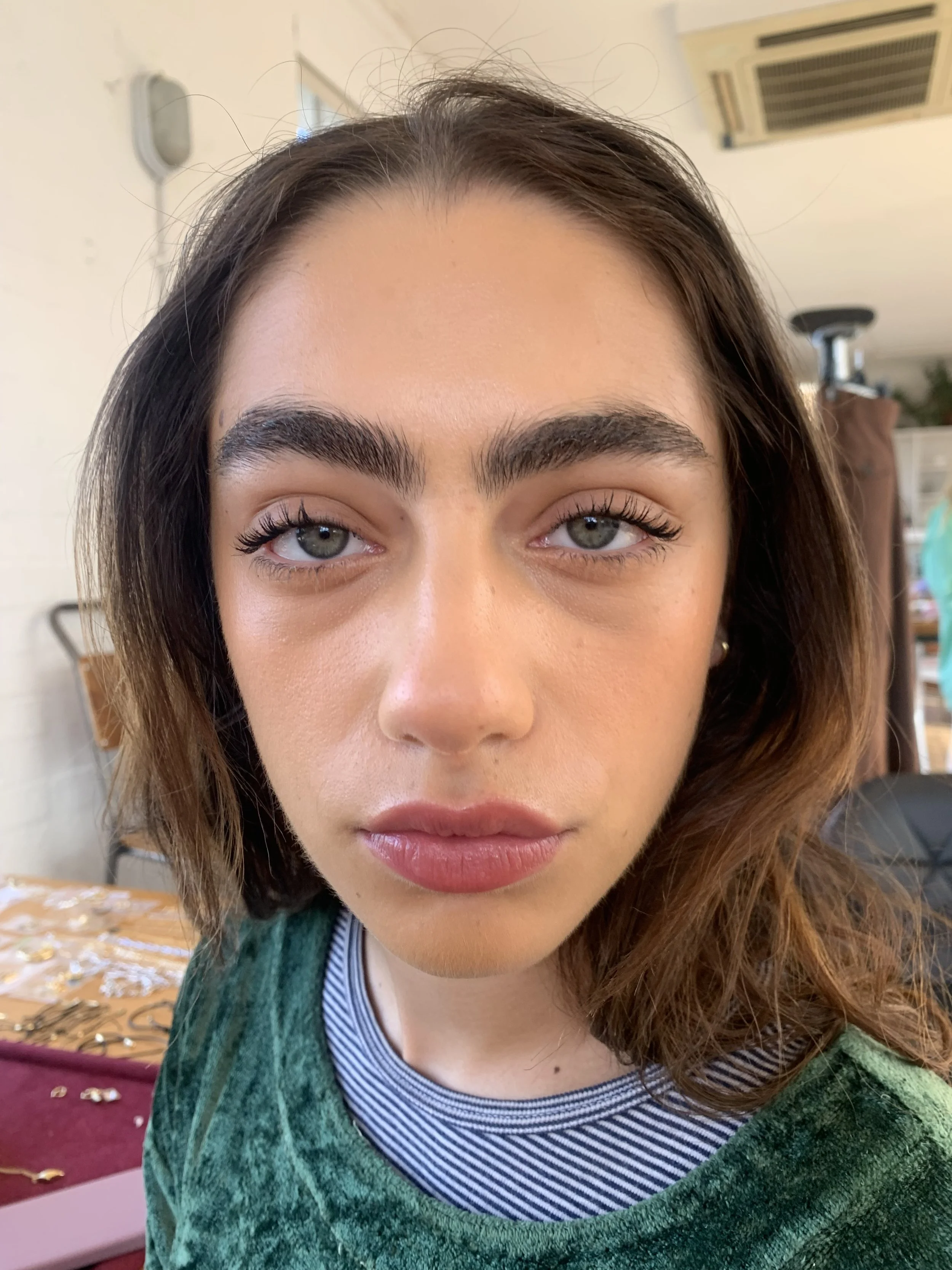 Close-up of a young woman with light skin, blue eyes, dark eyebrows, and wavy brown hair, wearing a green top with a striped neckline, indoors with a blurred background.