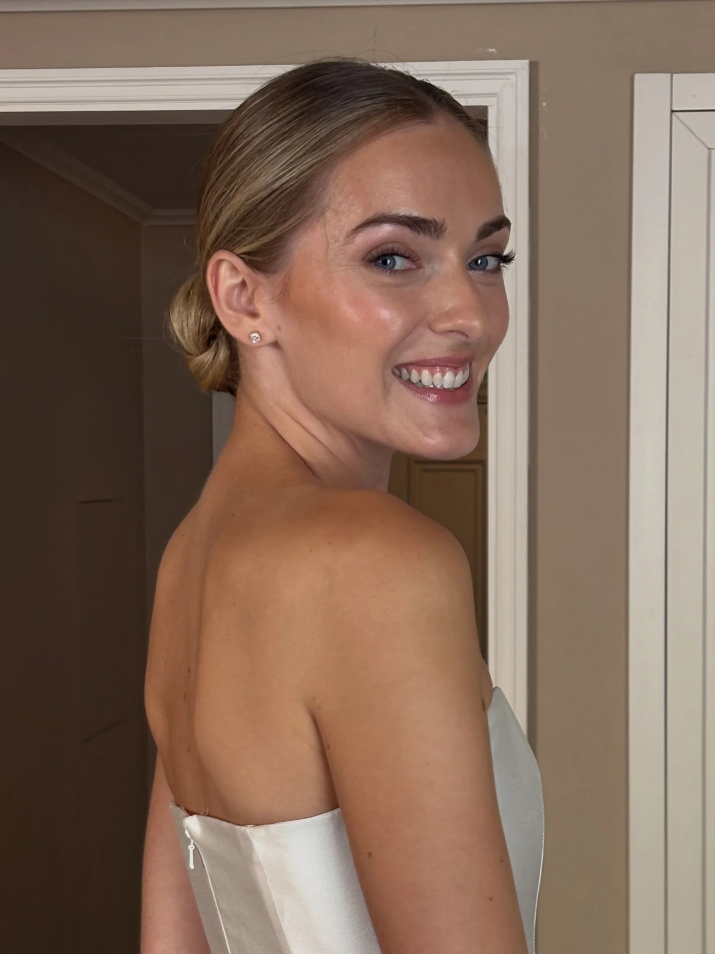 A woman with blonde hair styled in a low bun, wearing diamond stud earrings, smiling and looking over her shoulder. She is topless with a white satin piece of clothing around her waist, in a room with beige walls and white door trim.