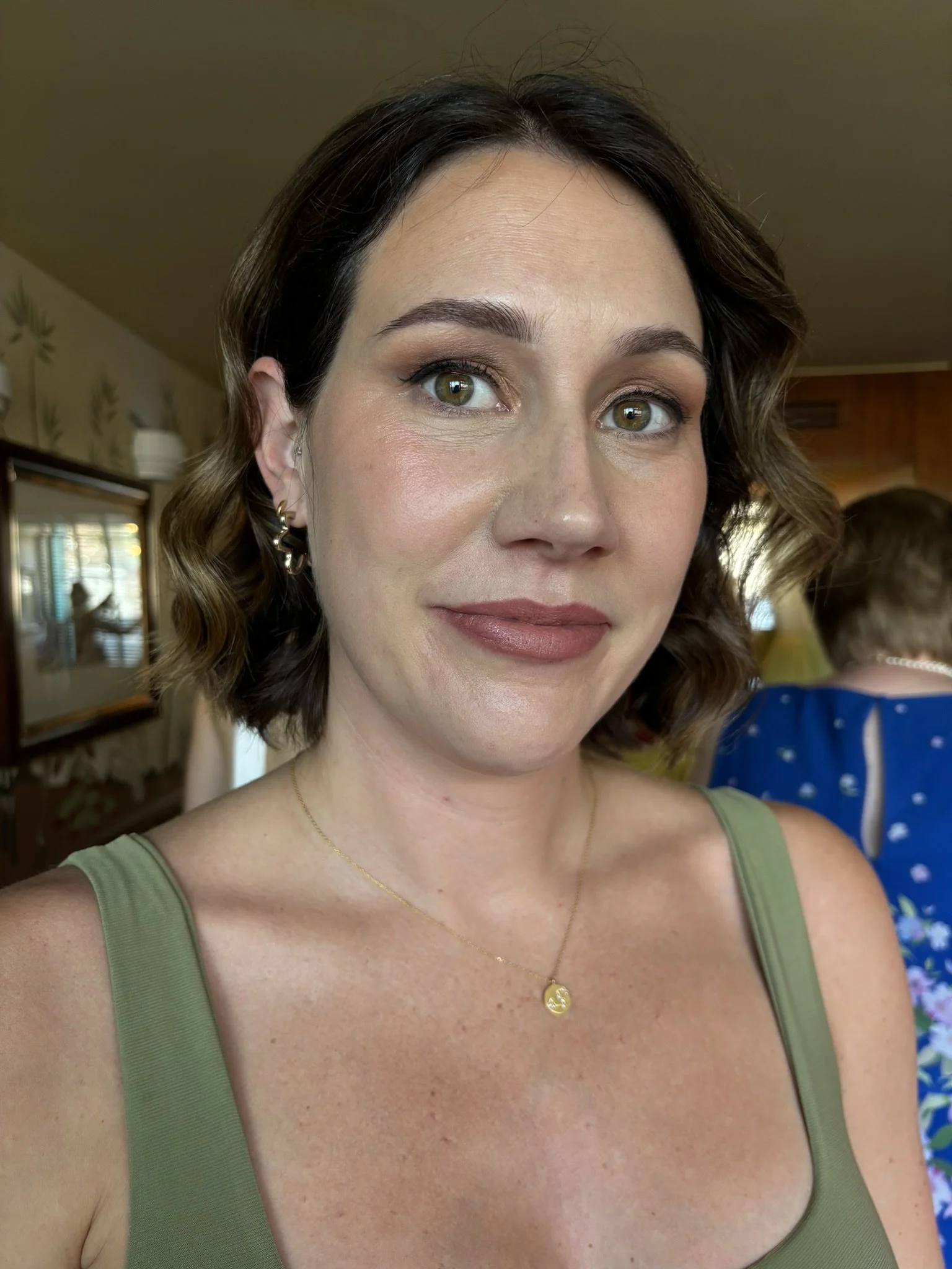 A woman with short wavy brown hair, green eyes, wearing a green tank top, a gold necklace, and earrings, smiling indoors.