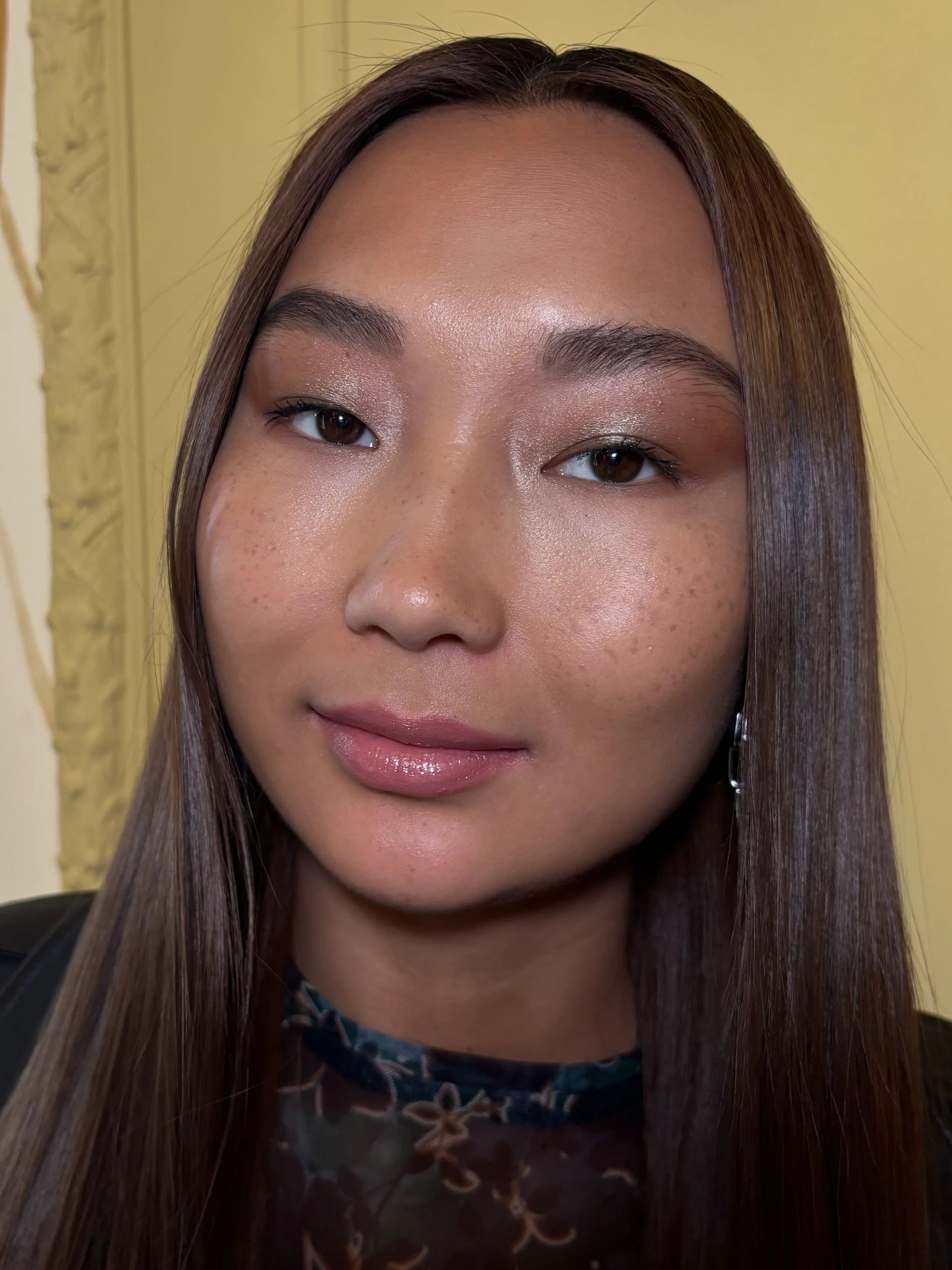 A young woman with long brown hair and brown eyes, smiling softly, wearing makeup with shiny lips and a floral-patterned top, standing in front of a yellow background.