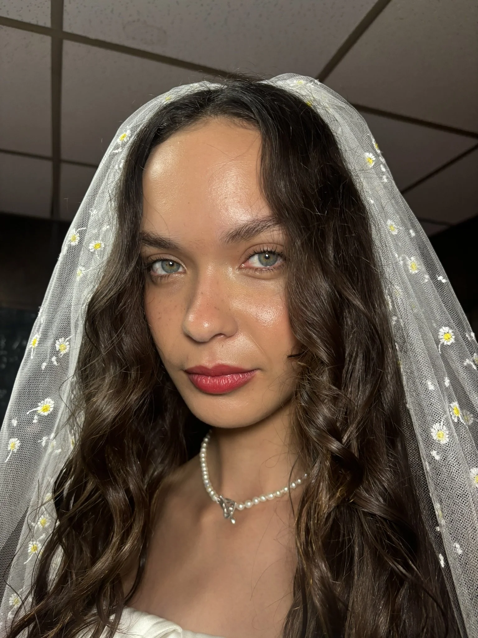 A woman with wavy brown hair wearing a sheer veil with daisies, pearl necklace, and makeup with red lipstick, looking directly at the camera.
