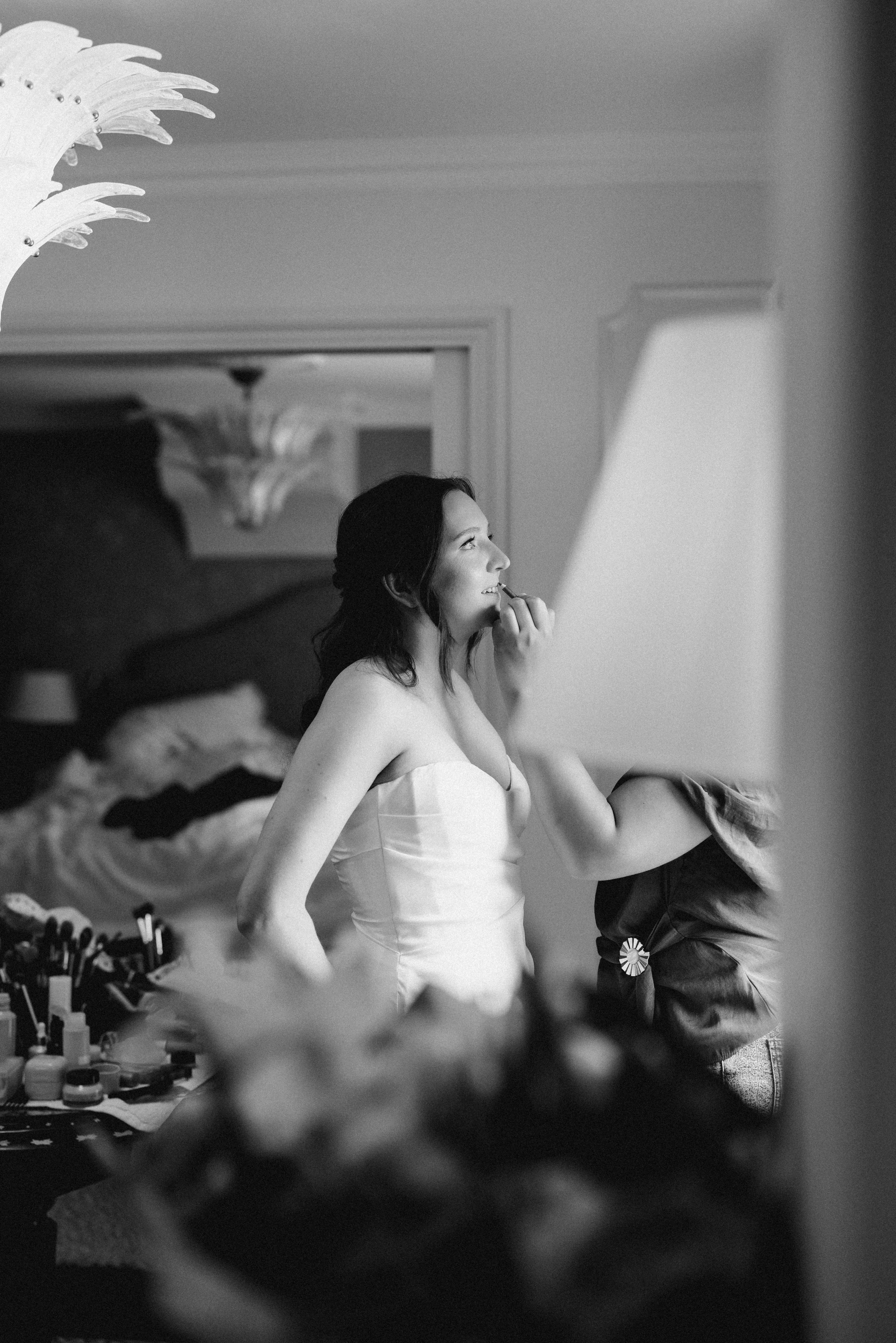 A woman applying makeup while sitting in a room with a mirror and makeup supplies on a table.