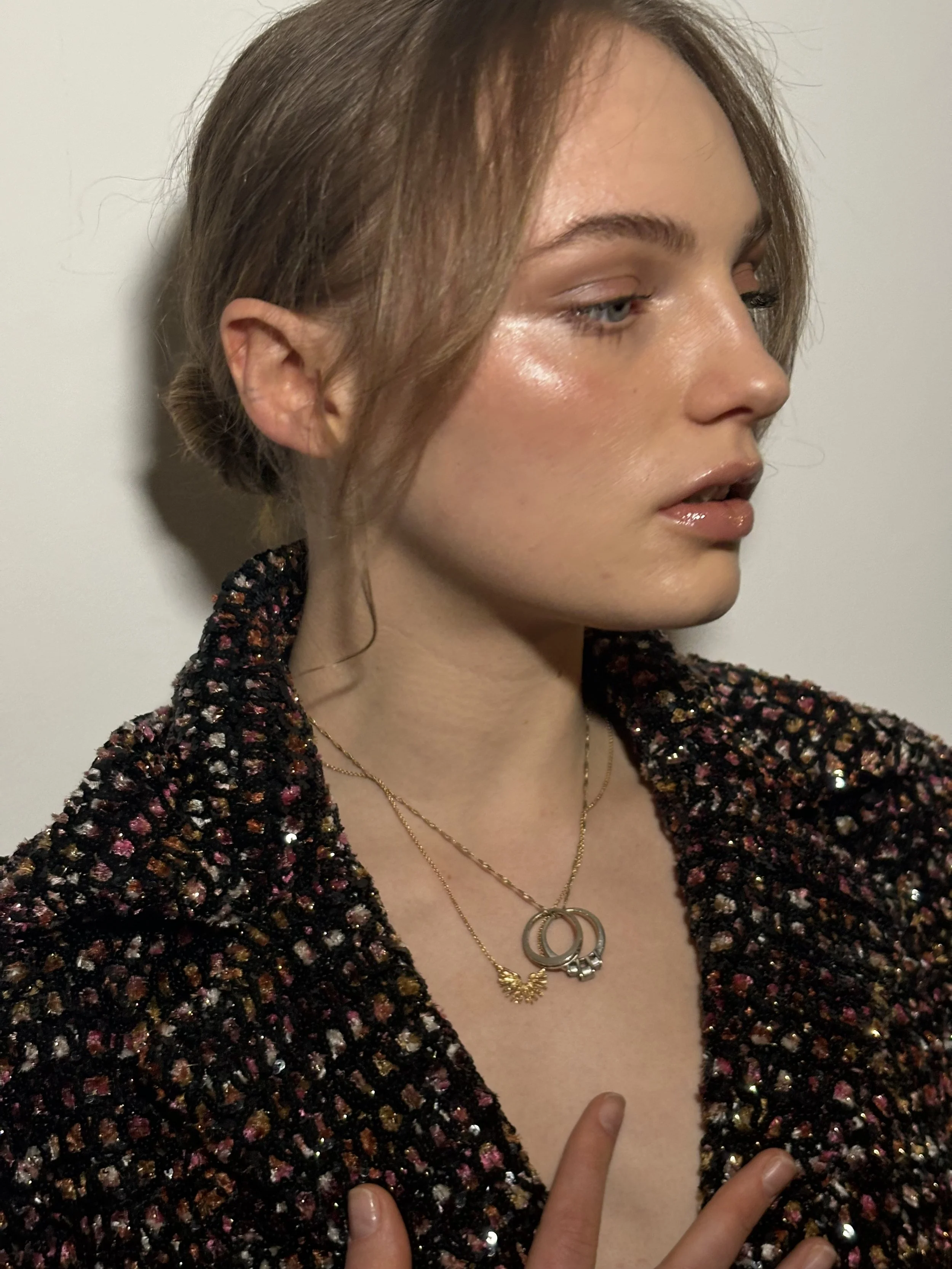 A young woman with light skin and reddish-brown hair styled in a loose updo looks to her left. She wears a multicolored, textured jacket and layered gold necklaces, including one with interlocking rings and another with a decorative wing charm.