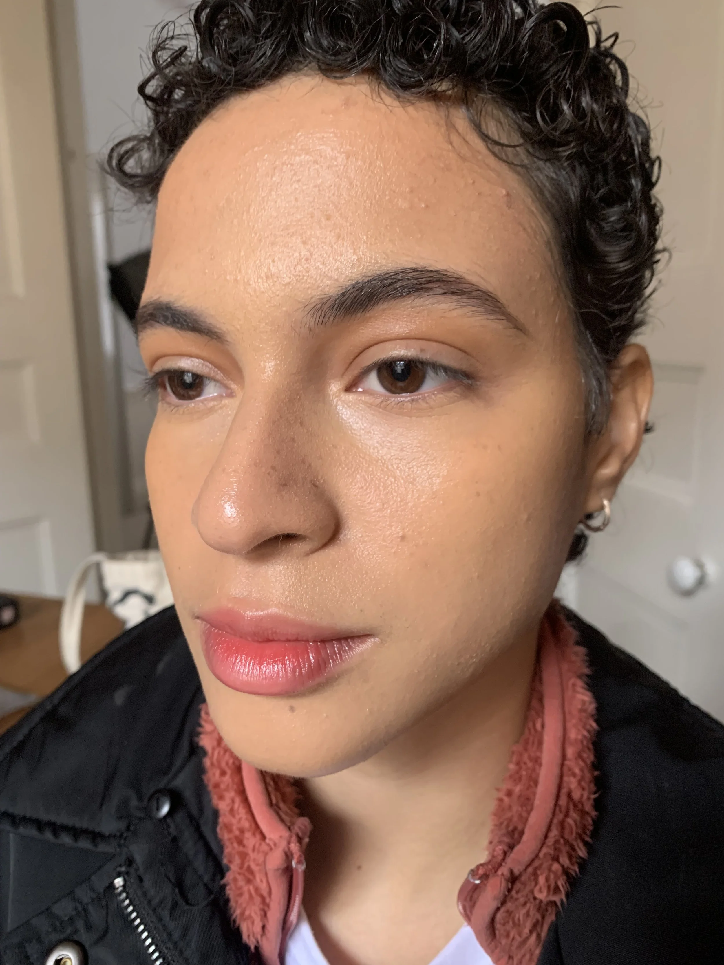 Close-up image of a person with curly hair, wearing a black jacket with a pinkish collar, and earrings, with a neutral facial expression.