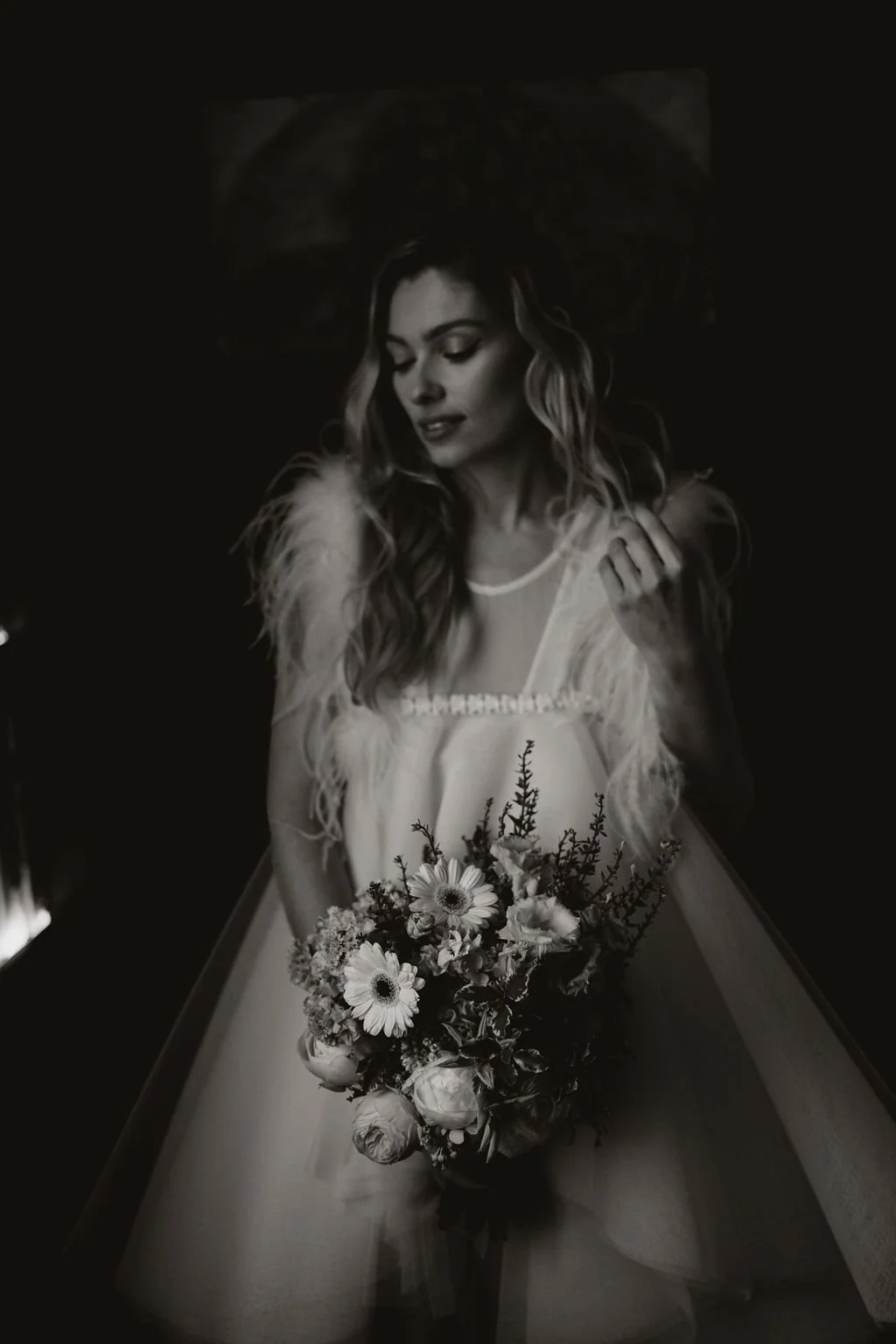 Black and white photo of a woman with wavy hair holding a bouquet of flowers, wearing a lacy silky dress with feathered sleeves, standing behind a table.