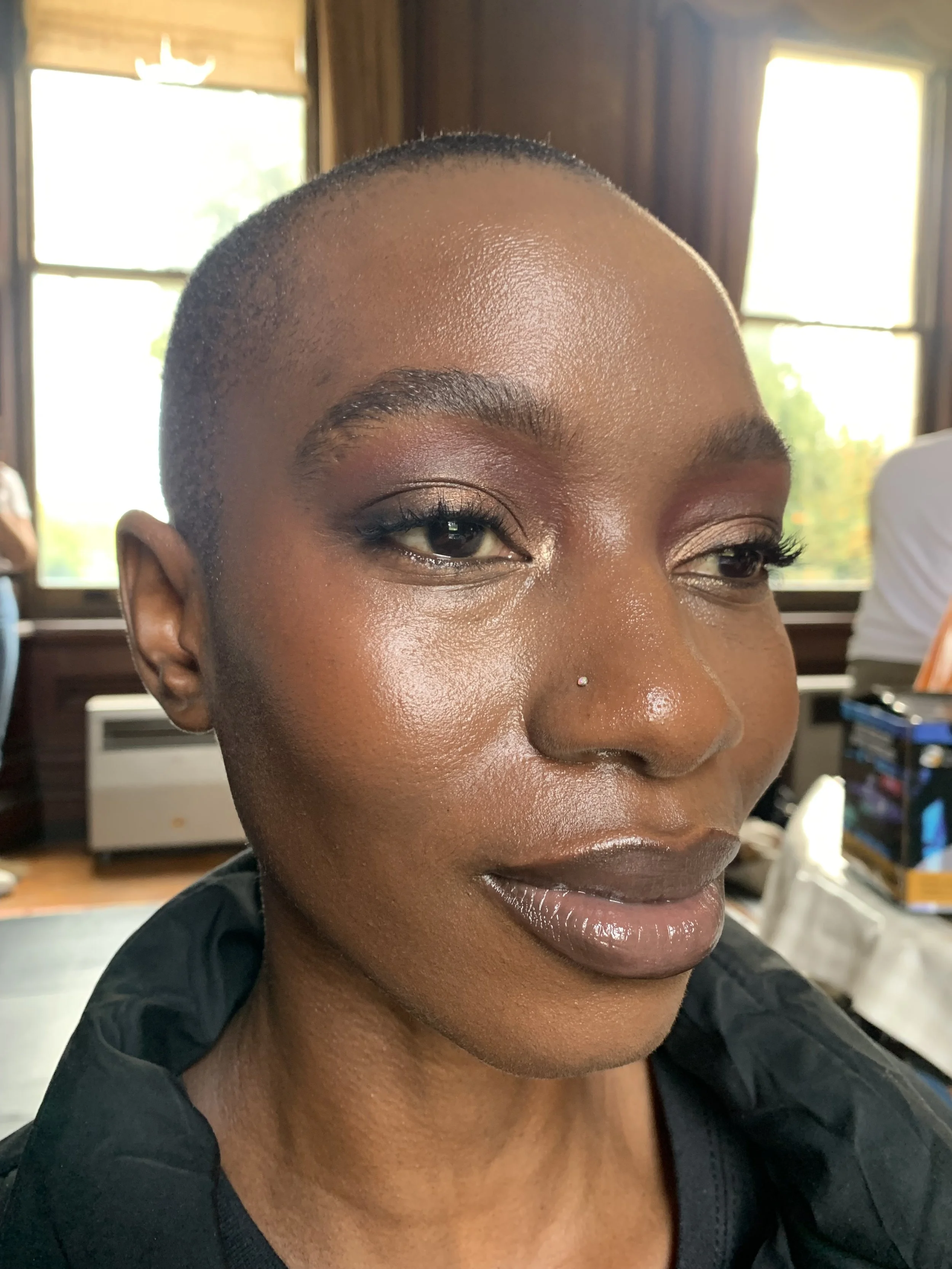Close-up of a woman with short hair, wearing makeup, and a small nose piercing, in a well-lit room with windows.