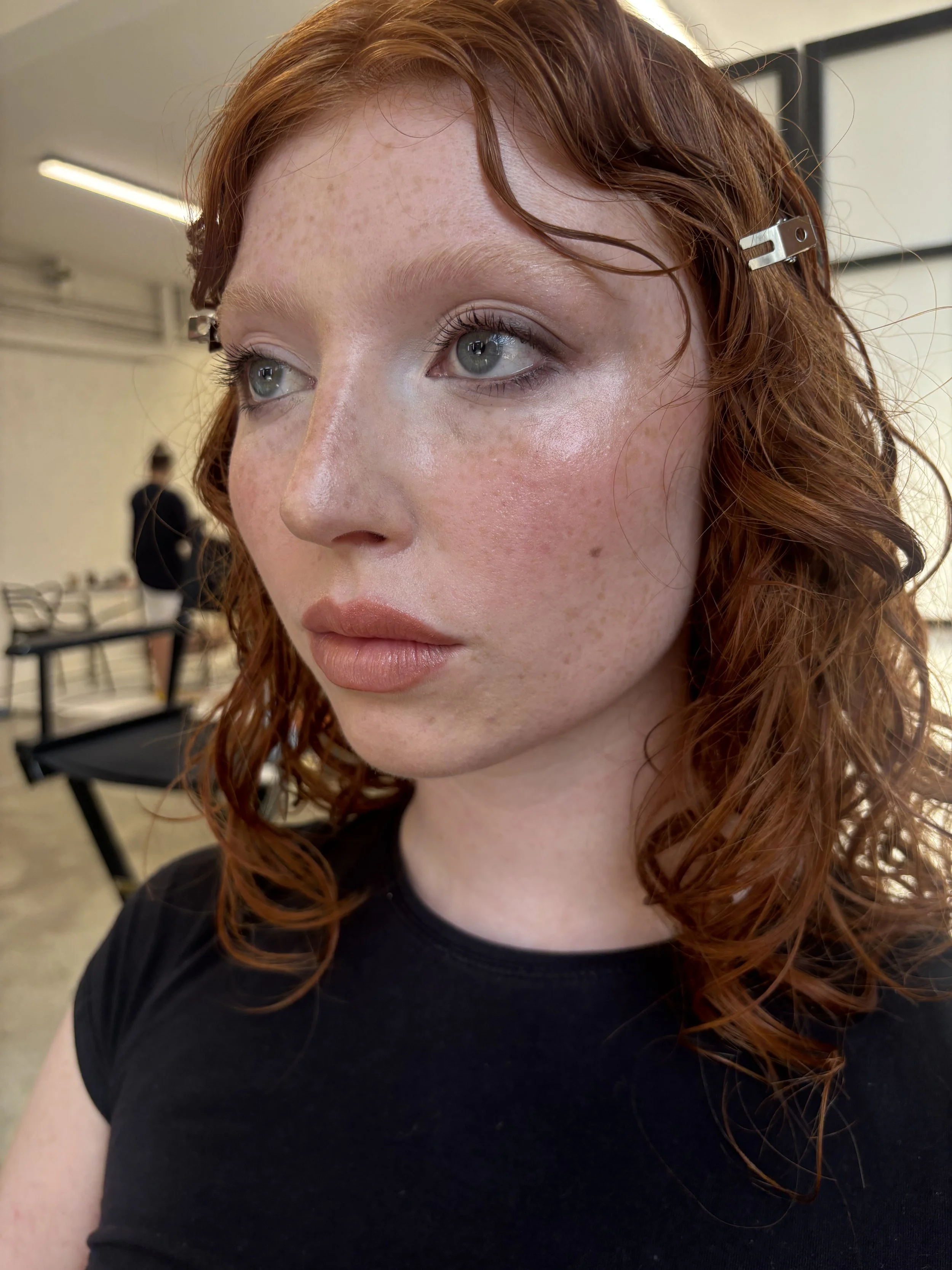 Close-up of a woman with red curly hair held back with metal hair clips, light makeup, and wearing a black shirt, in an indoor setting with blurred background.