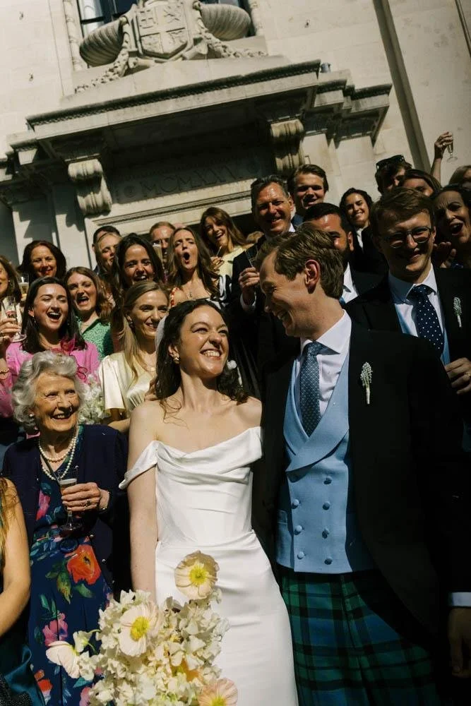 A wedding celebration outdoors with the bride and groom smiling at each other, surrounded by laughing guests, with a historic building and a statue in the background.