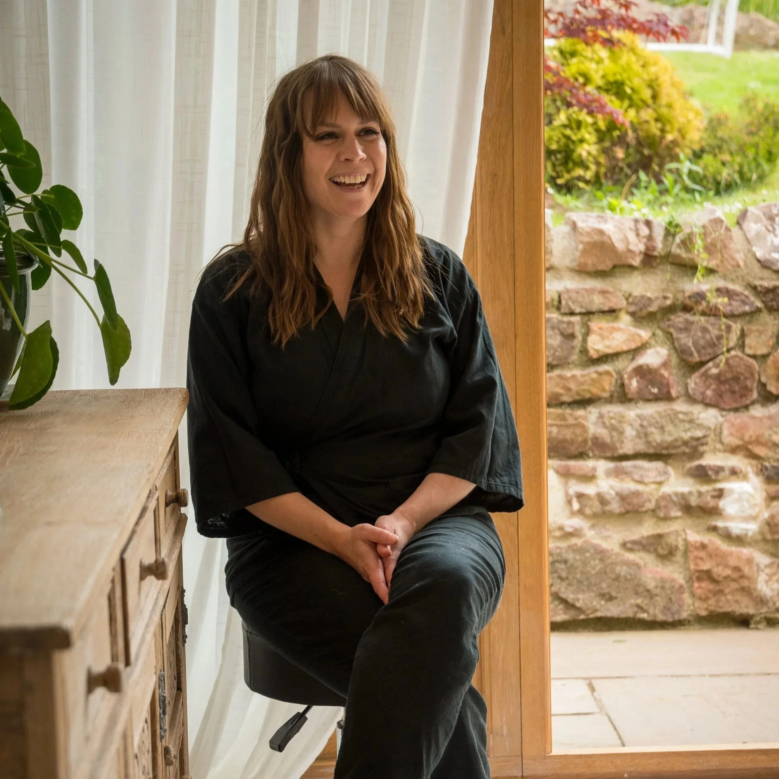 A smiling woman with long reddish-brown hair, wearing a black shirt, is sitting indoors near a door with a view of a garden with vibrant red and yellow foliage outside.