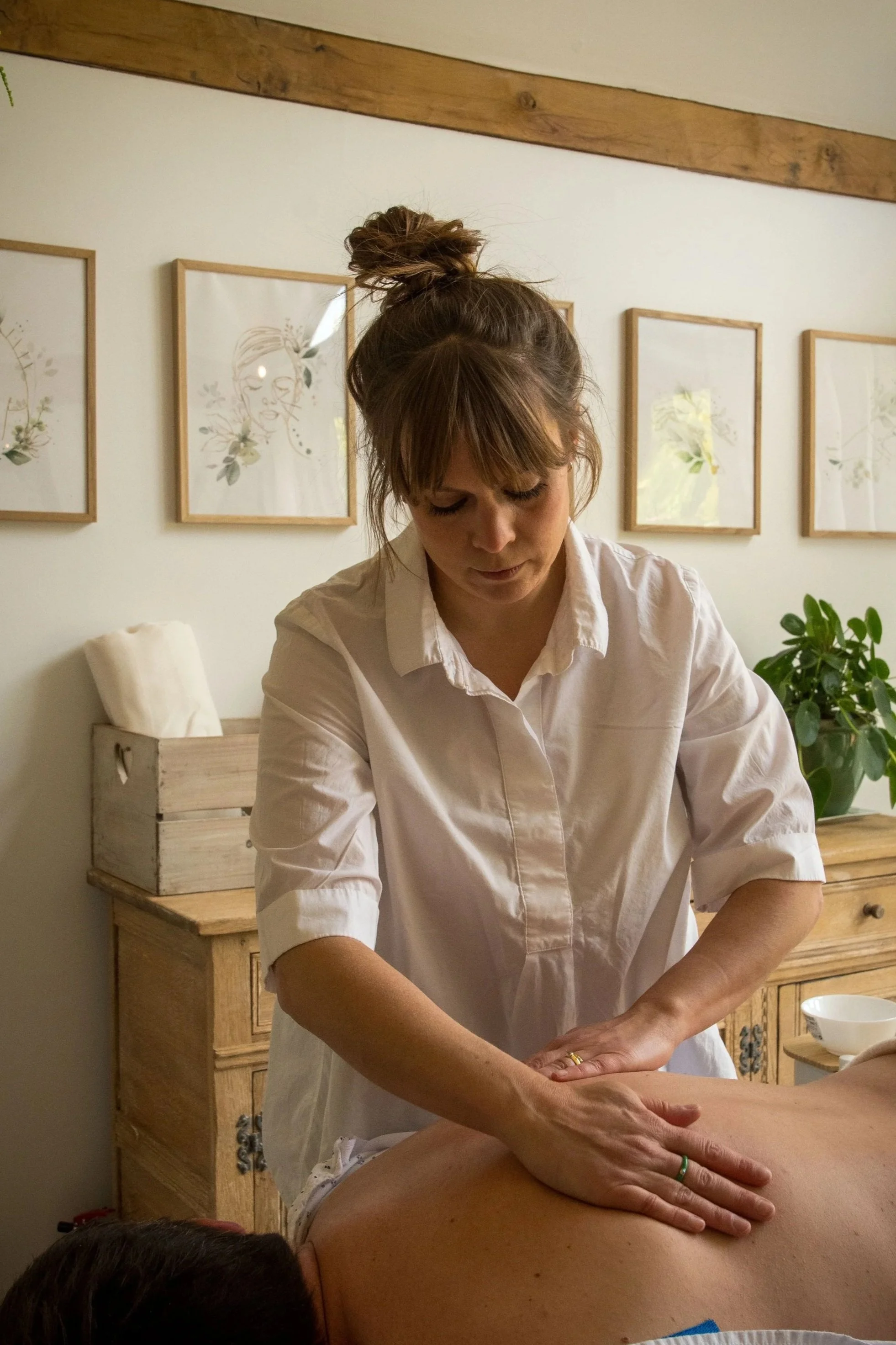 A woman giving a massage to a person lying face down on a massage table, with framed art on the wall behind them and a wooden cabinet nearby.