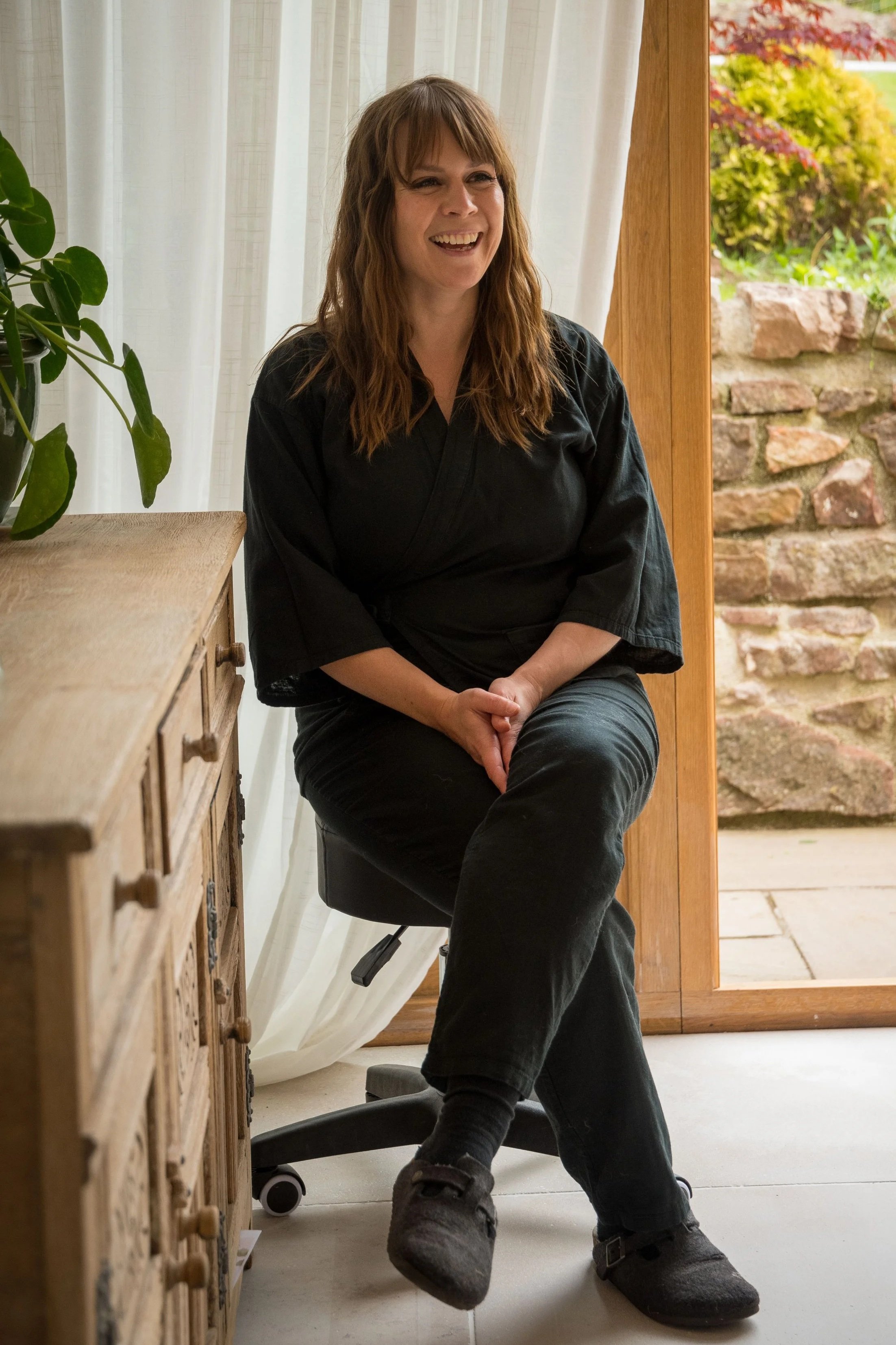 A woman with brown hair sitting on an office chair, smiling, next to a wooden cabinet and a window with white curtains, with a garden visible outside.