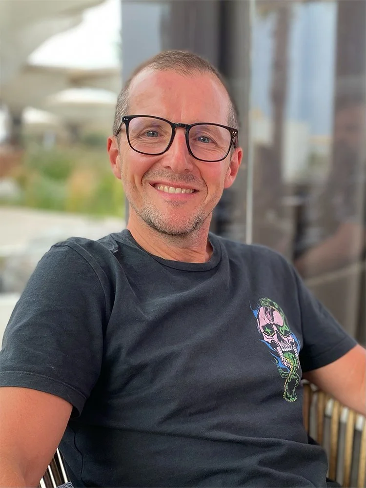 A smiling man wearing glasses and a black T-shirt with a colorful skull and snake graphic, sitting indoors with a blurred background.