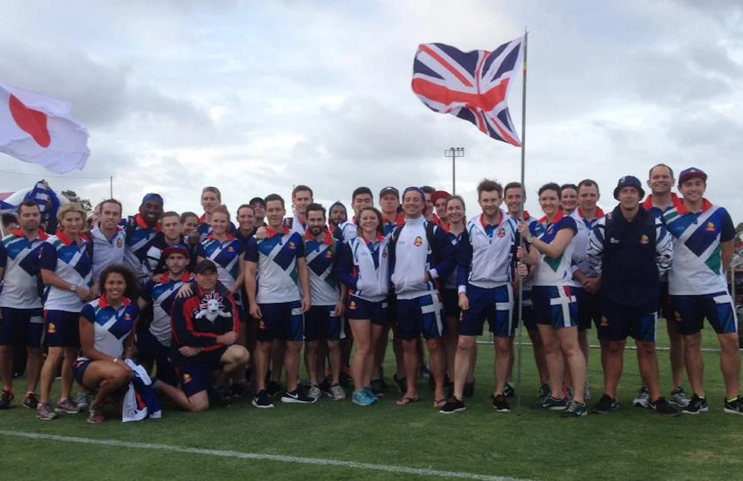 Group of athletes and staff standing on a field, wearing team uniforms with blue, white, and green patterns, holding flags of the United Kingdom and Greece, under cloudy sky.
