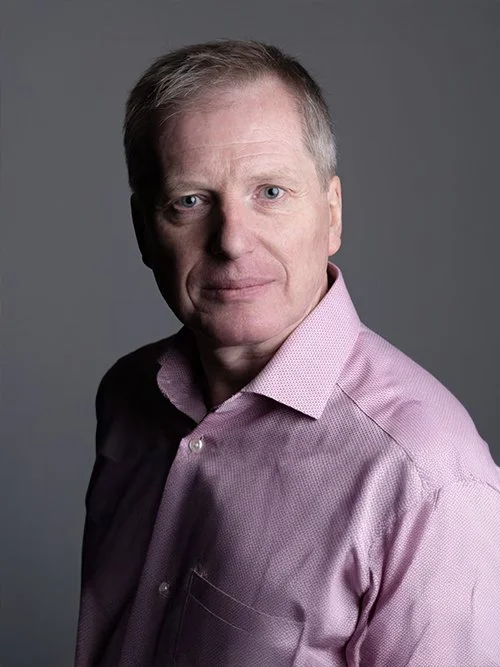 A middle-aged man with short light brown hair and blue eyes, wearing a pink dress shirt, looking at the camera against a neutral gray background.