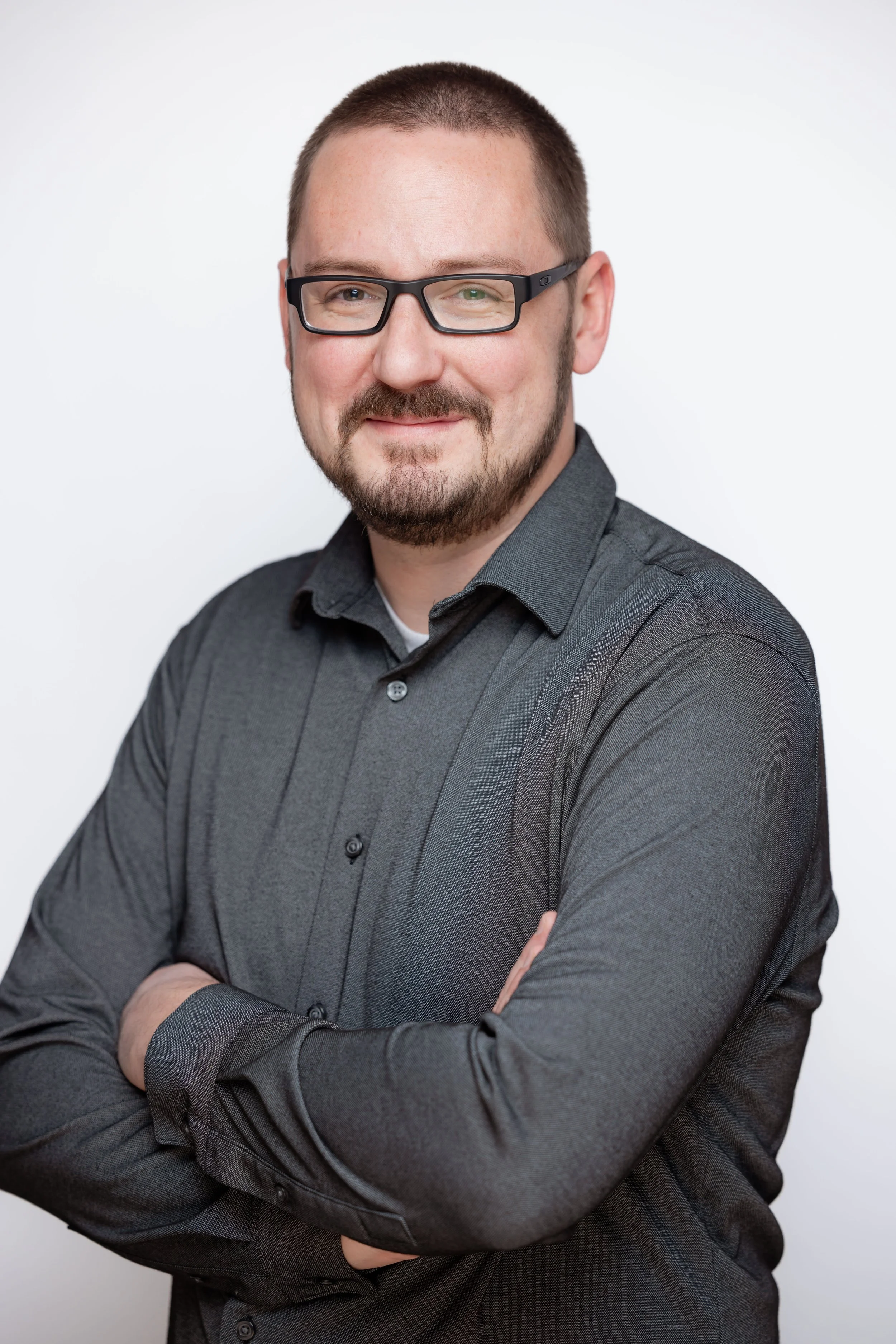 Headshot of a man with glasses and a beard, wearing a dark gray button-up shirt with arms crossed, smiling against a plain white background.