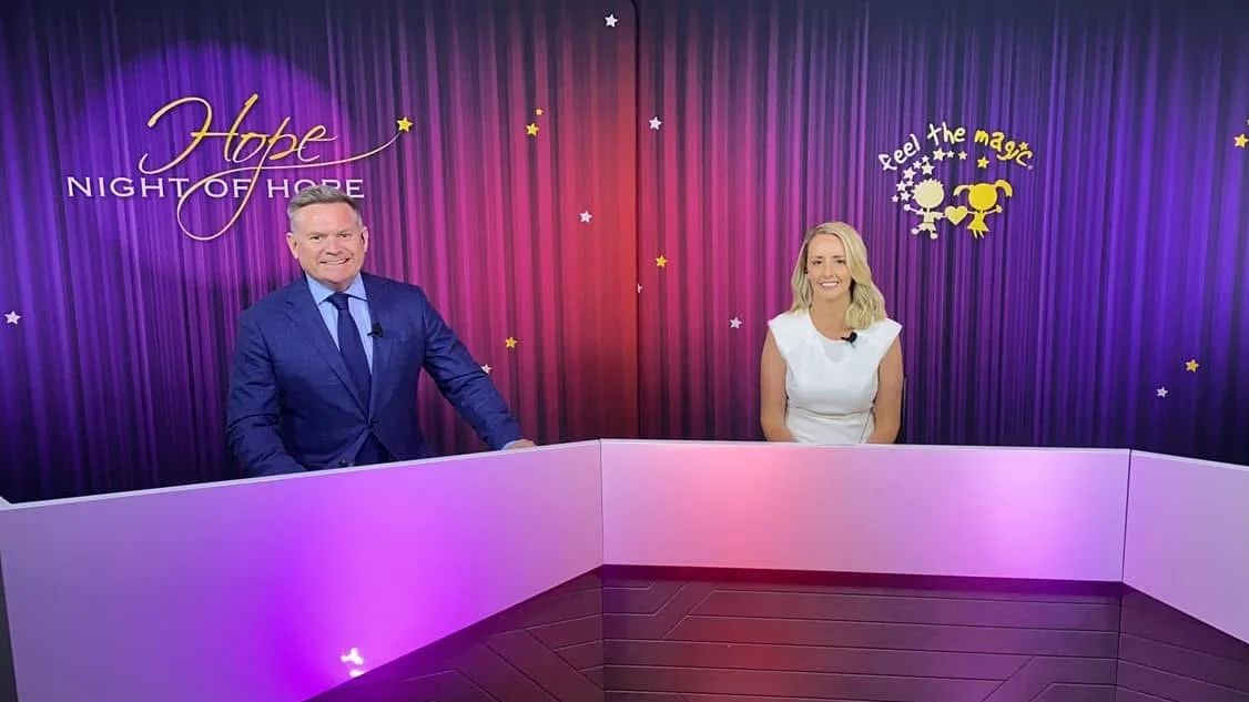 Studio set with two hosts, a man in a blue suit and a woman in a white dress, sitting behind a curved white desk with purple lighting. Behind them, a purple curtain with stars, and signs that say 'Hope Night of Hope' and 'Feel the magic' with illustrations of children holding hands.