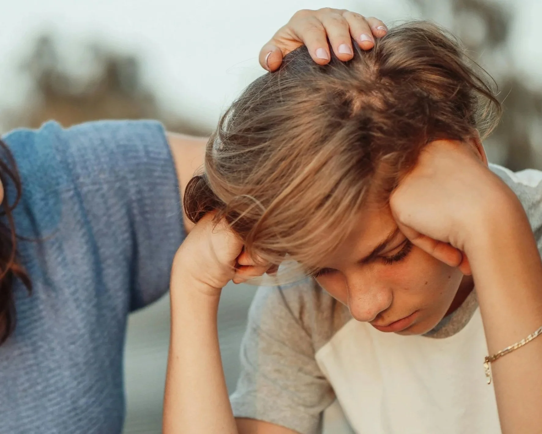 A young person with brown hair and light skin appears distressed, with their head bowed and eyes closed, holding their forehead with one hand while an adult gently touches their head.