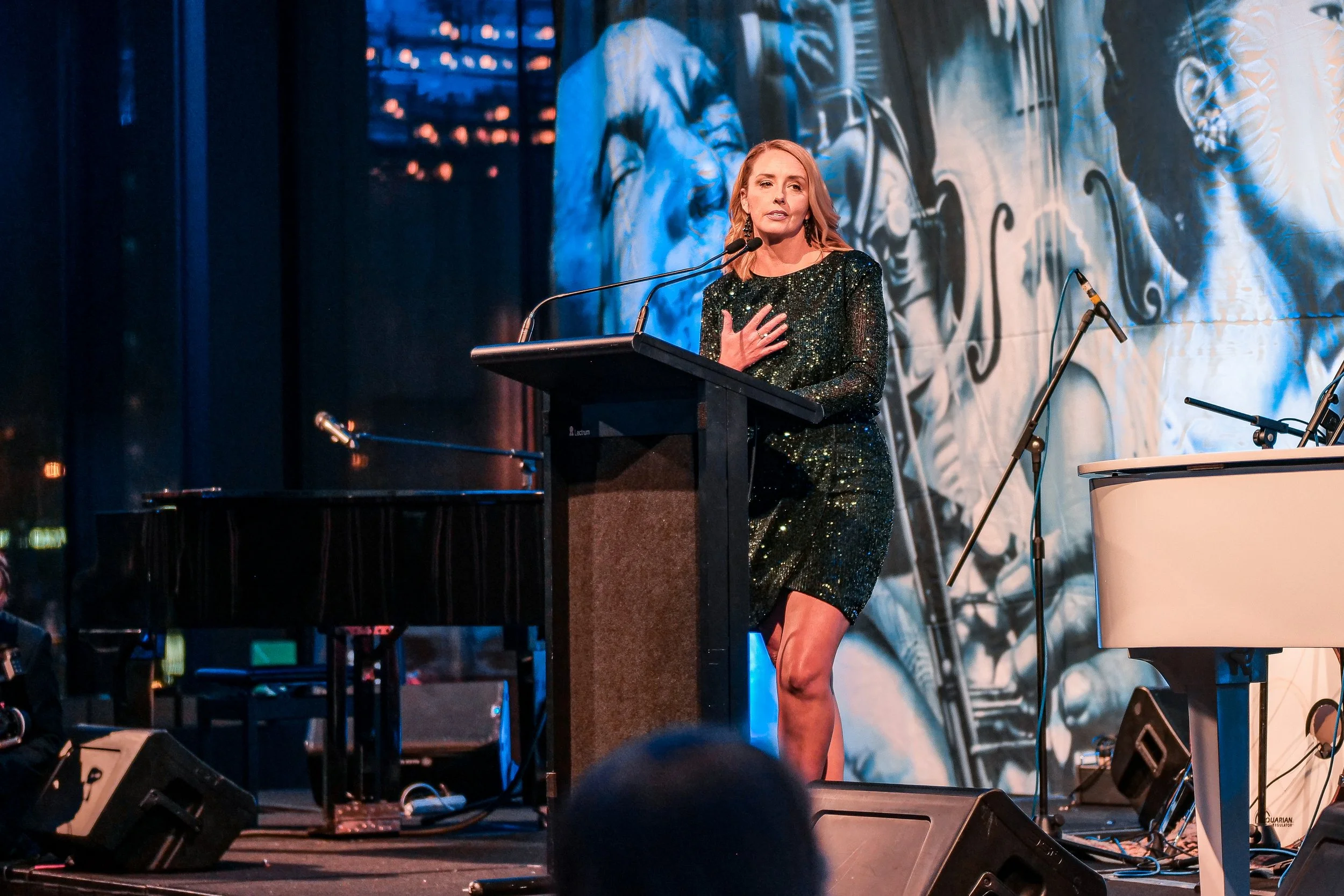 A woman in a sparkly black dress giving a speech at a podium on a stage with a large mural in the background.