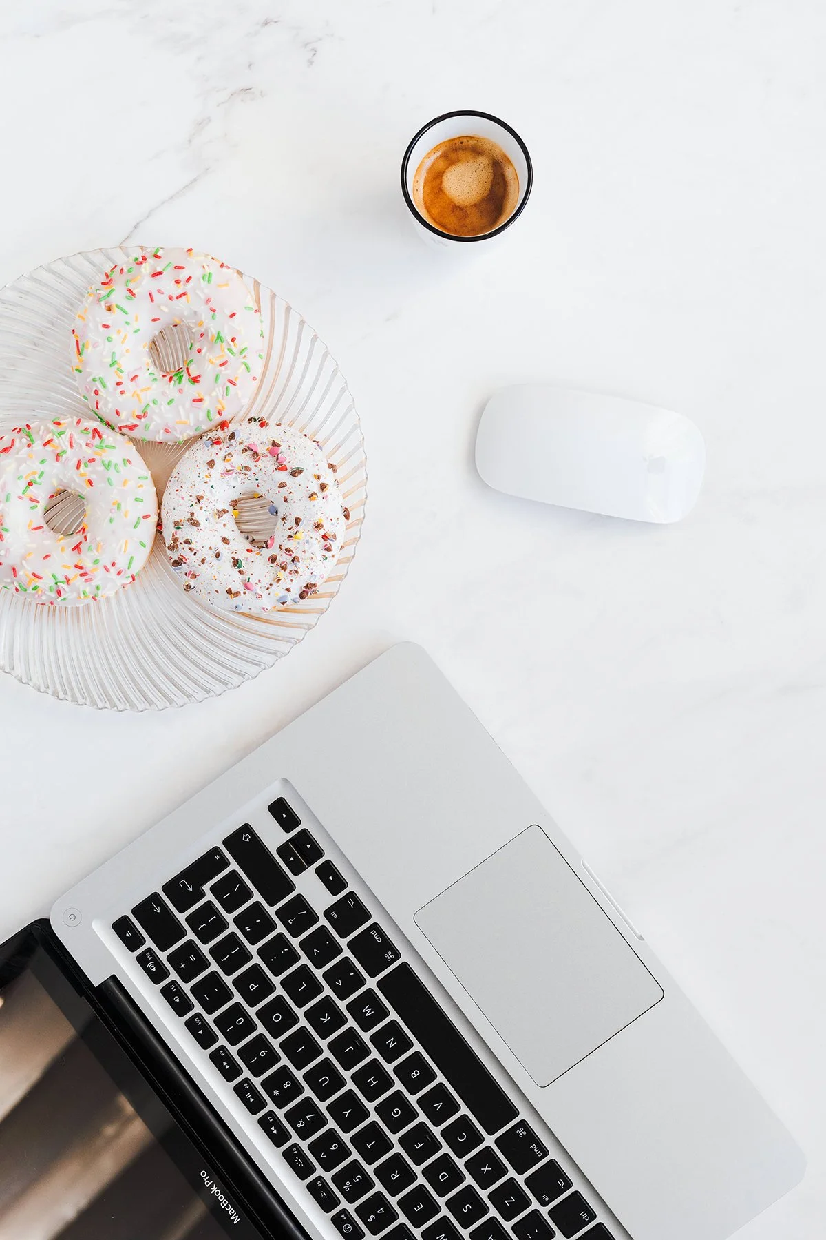 A workspace with a silver laptop, a white computer mouse, a glass of coffee, and a plate with three donuts topped with colorful sprinkles and icing on a white marble surface.