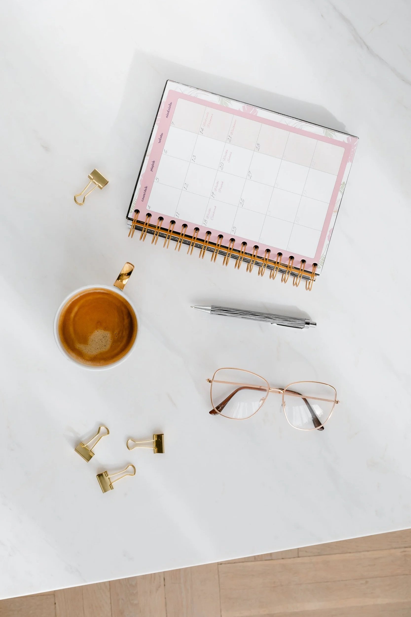 A white desk with a notebook, coffee mug, pen, glasses, and gold binder clips.