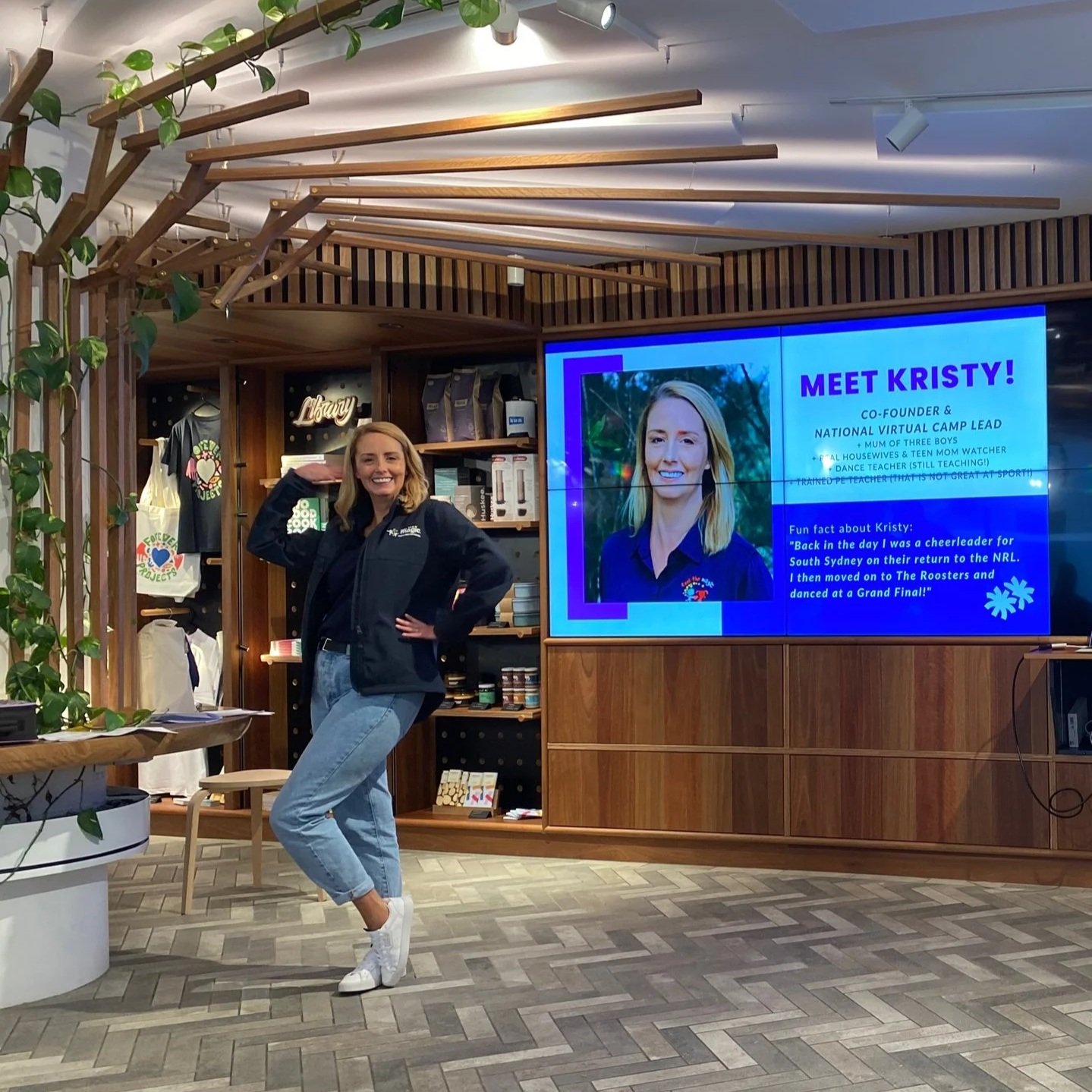 A woman standing in a retail store next to a large digital screen displaying a profile of Kristy, a co-founder and camp lead, with her photo and fun facts. She is smiling, wearing a black jacket, jeans, and white sneakers. The store has wooden accents and merchandise on shelves.