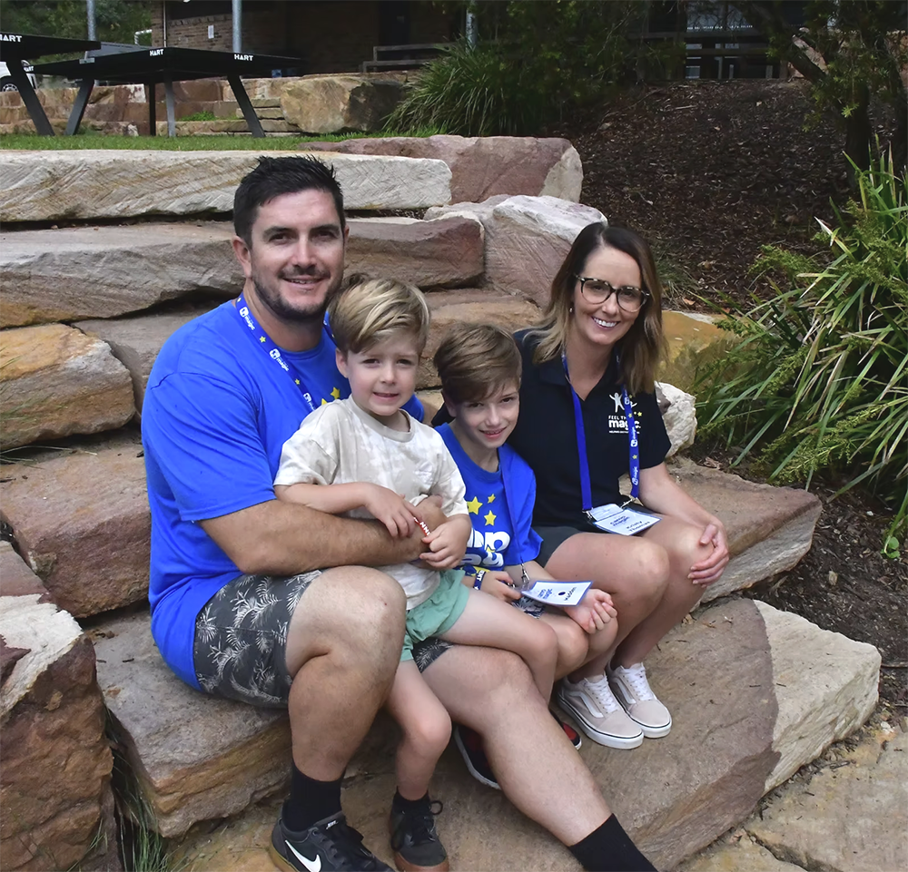 A family of four sitting on a stone staircase outdoors, smiling at the camera. The father is wearing a blue shirt and patterned shorts, holding a young boy. The mother is wearing glasses and a black shirt, holding a young girl. The family appears happy and relaxed.