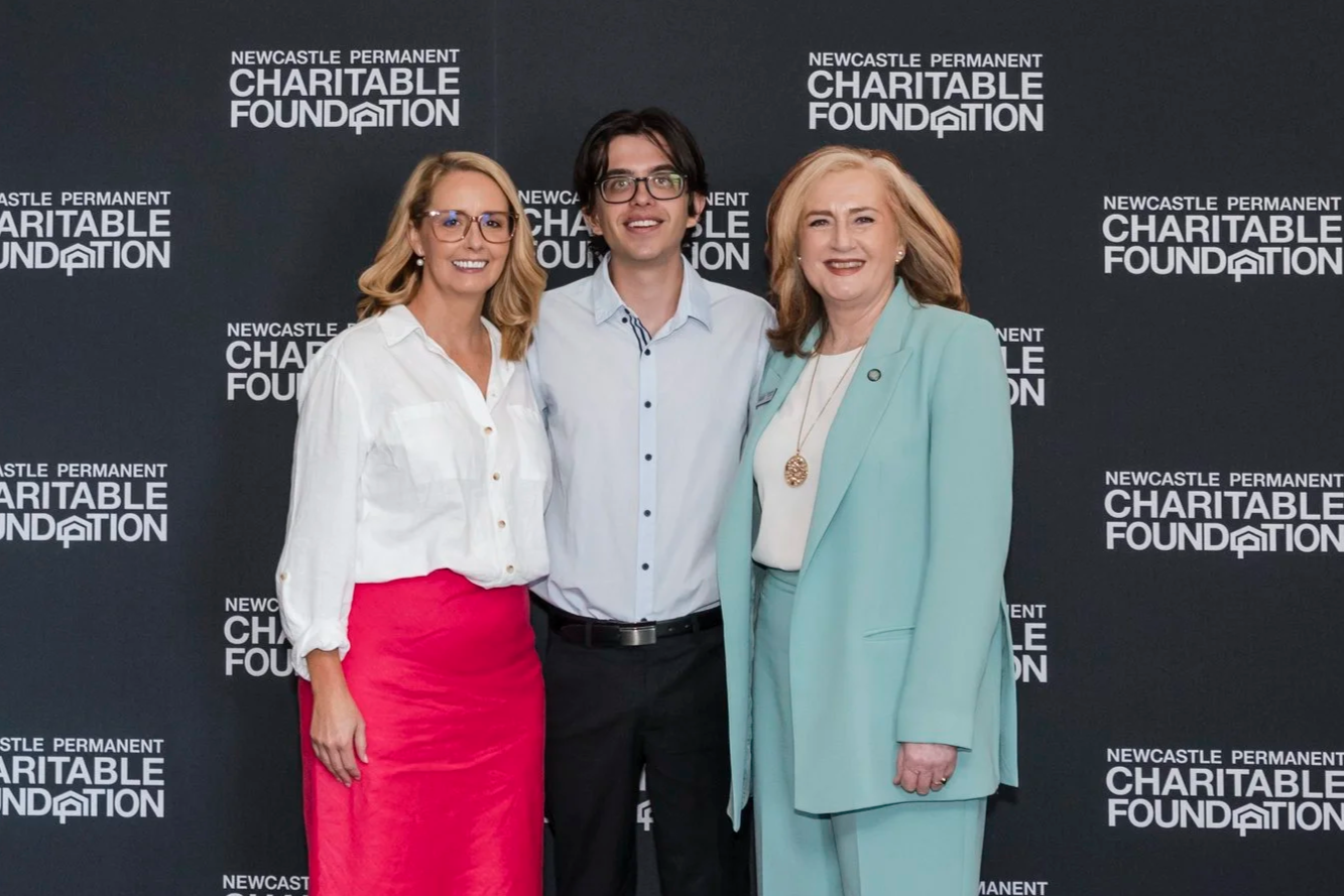 Three people standing together smiling at an event for the Newcastle Permanent Charitable Foundation, against a backdrop with the foundation's logo.
