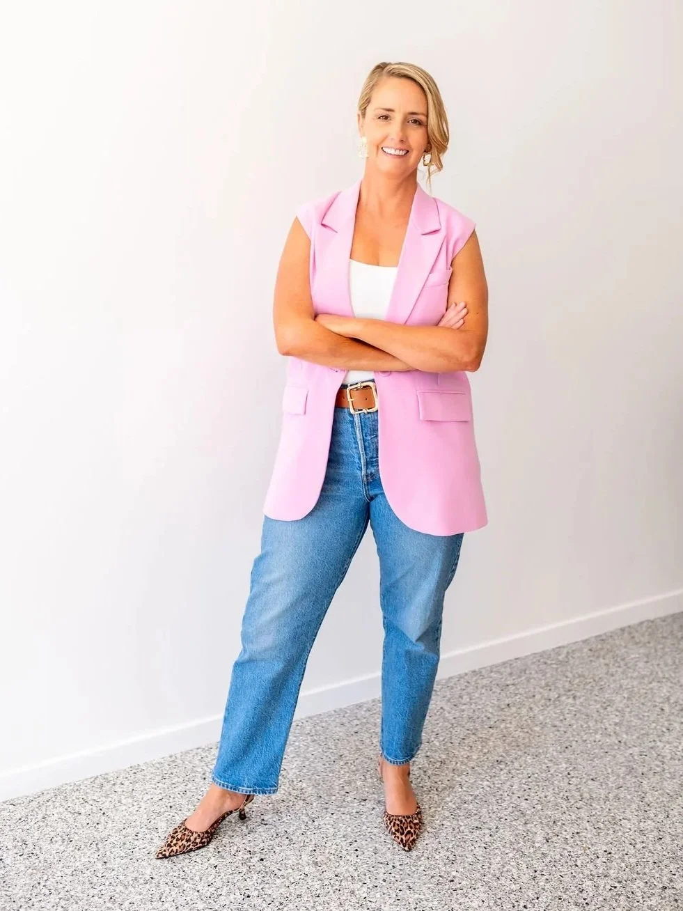 A woman with blonde hair, wearing a pink sleeveless blazer, white top, blue jeans, leopard print heels, smiling with arms crossed, standing against a plain white wall.