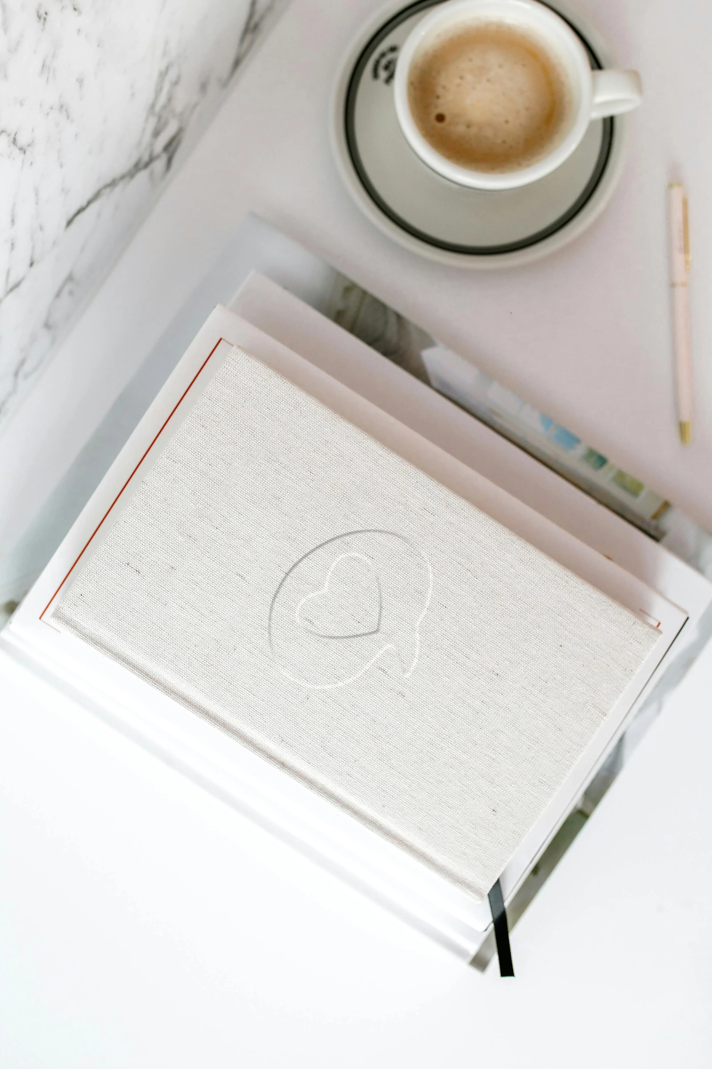 A white book with a heart and speech bubble embossed on the cover, placed on a white desk next to a cup of coffee and a pen.