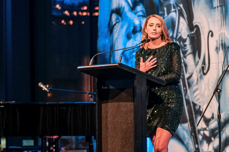 A woman with blonde hair standing at a podium giving a speech, with a microphone in front of her, hand on her chest, wearing a sparkly black dress, in front of a blue and black artistic backdrop.