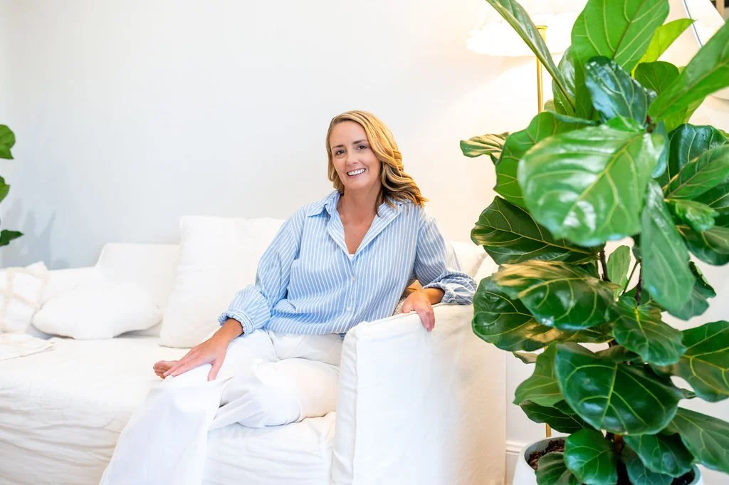A woman with blonde hair, wearing a blue and white striped shirt, sits on a white couch, smiling in a bright, modern living room with large green houseplants.