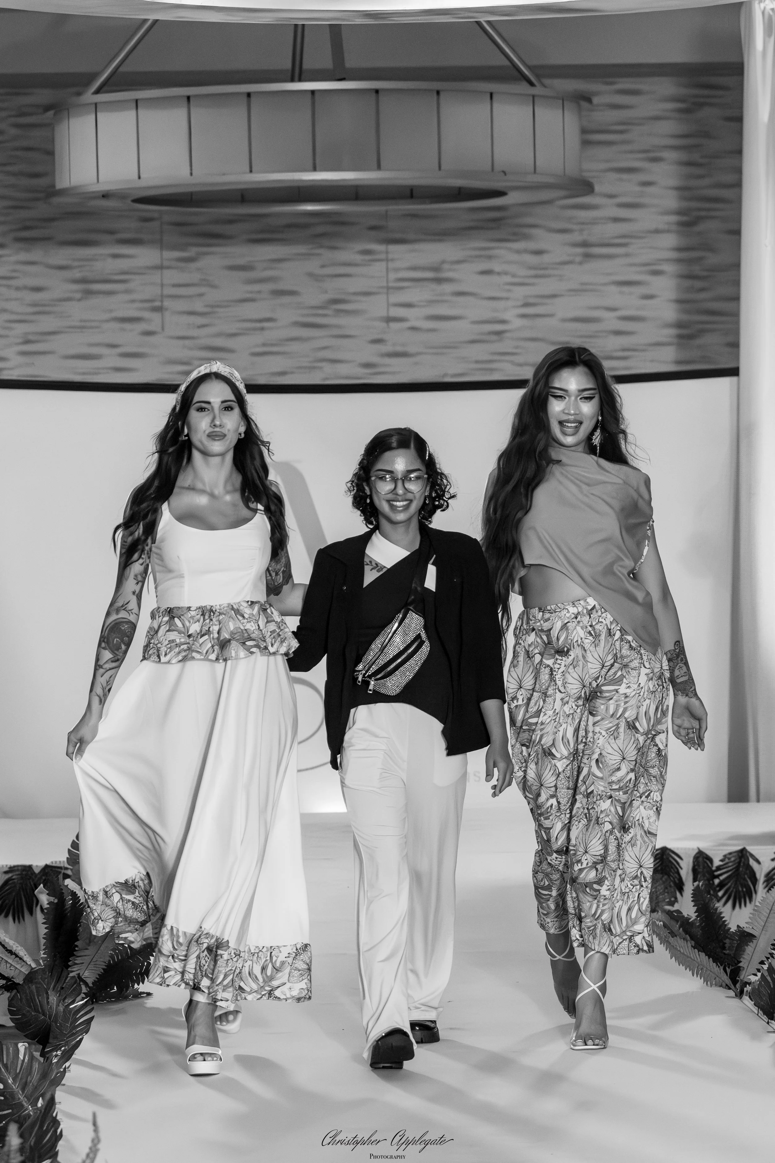 Black and white photo of three women walking down a runway, showing fashion outfits during a fashion show. The background features water imagery.