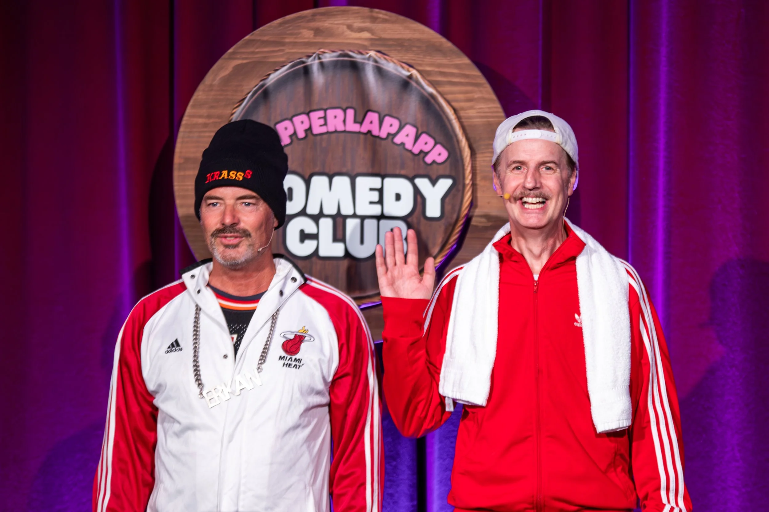 Zwei Männer auf einer Comedy-Bühne, einer hebt die Hand, im Hintergrund ein Schild mit dem Text "Papperlapapp Comedy Club".