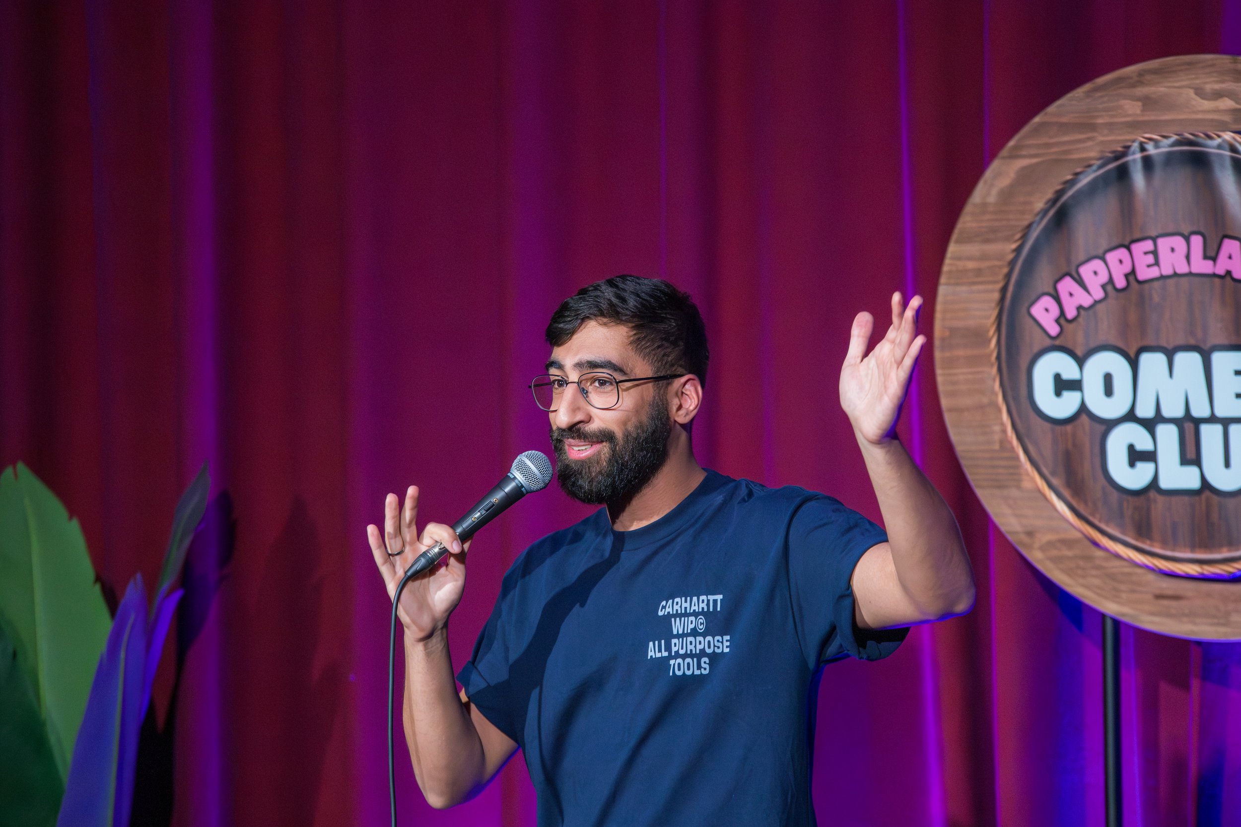 Mann steht auf einer Bühne, hält ein Mikrofon, trägt ein blaues T-Shirt mit Aufdruck und winkt mit der Hand. Im Hintergrund ist ein Vorhang und ein rundes Schild mit der Aufschrift "PAPPERLAND COMEDY CLUB".