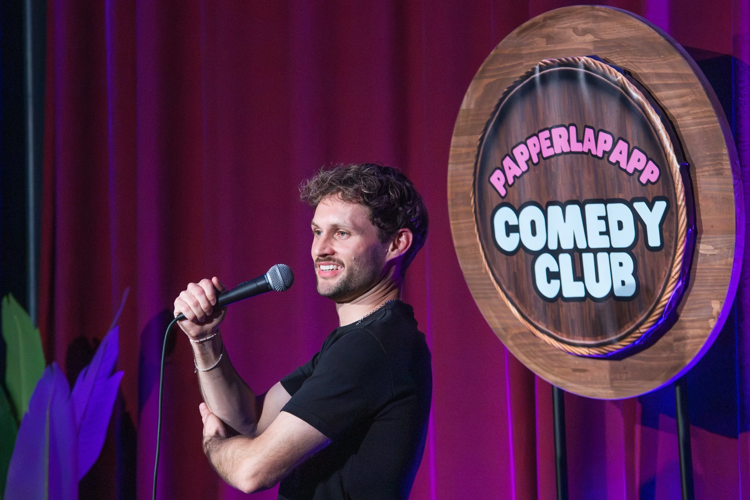 Ein Mann steht auf einer Bühne des Paperlapapp Comedy Clubs, hält ein Mikrofon in der Hand und spricht. Im Hintergrund ist ein großes rundes Schild mit dem Schriftzug 'PAPERLAPAPP COMEDY CLUB' zu sehen, beleuchtet mit buntem Licht.