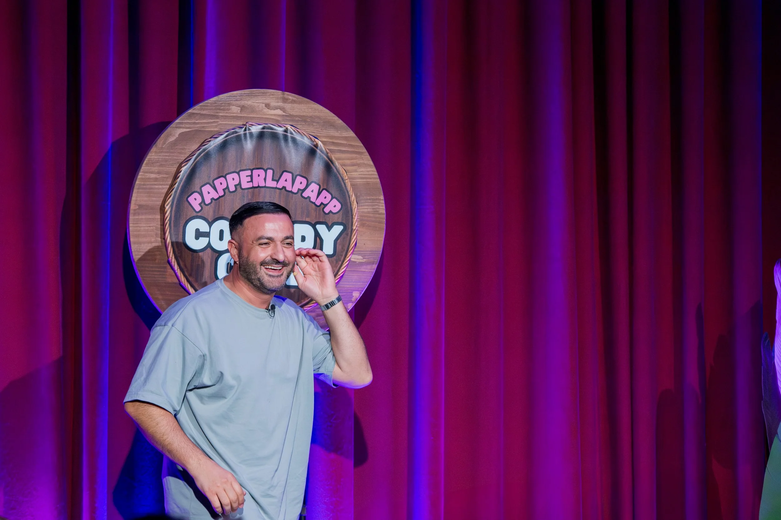 Ein Mann lächelt auf einer Bühne vor einem roten Vorhang, hinter ihm ein großes Schild mit der Aufschrift "PAPPERLAPAPP COMEDY".