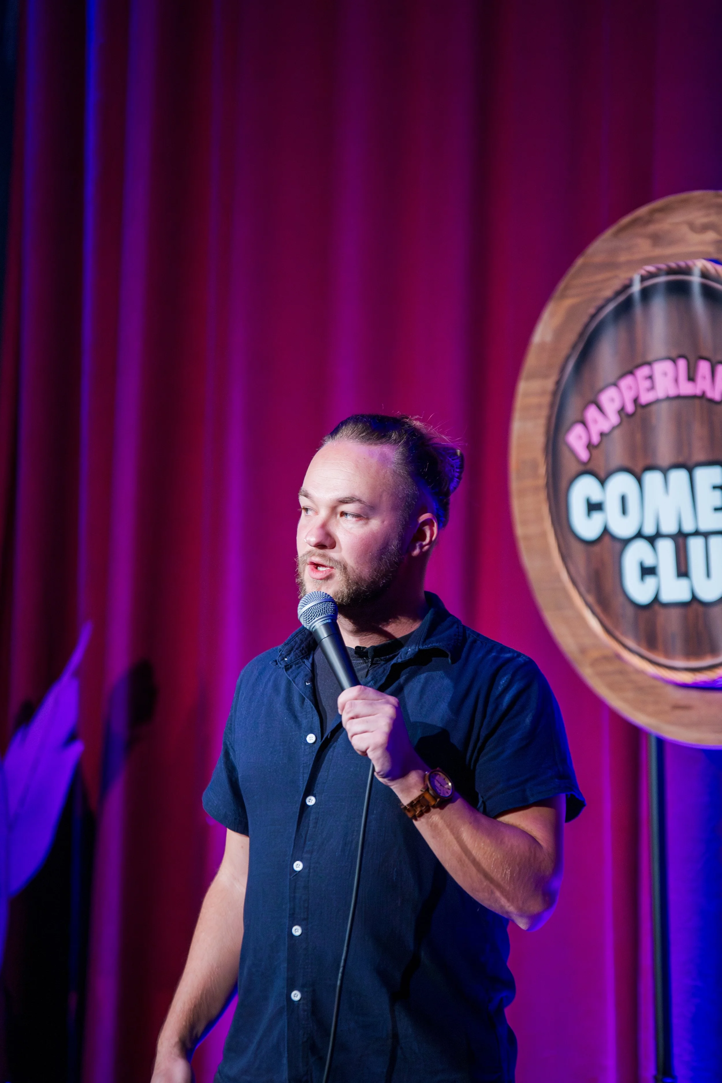 Mann steht auf einer Comedy-Bühne mit rotem Vorhang im Hintergrund, hält ein Mikrofon in der Hand und trägt eine dunkle Hemdjacke. Ein rundes Schild mit der Aufschrift 'PAPERLAND COMEDY CLUB' ist im Hintergrund sichtbar.
